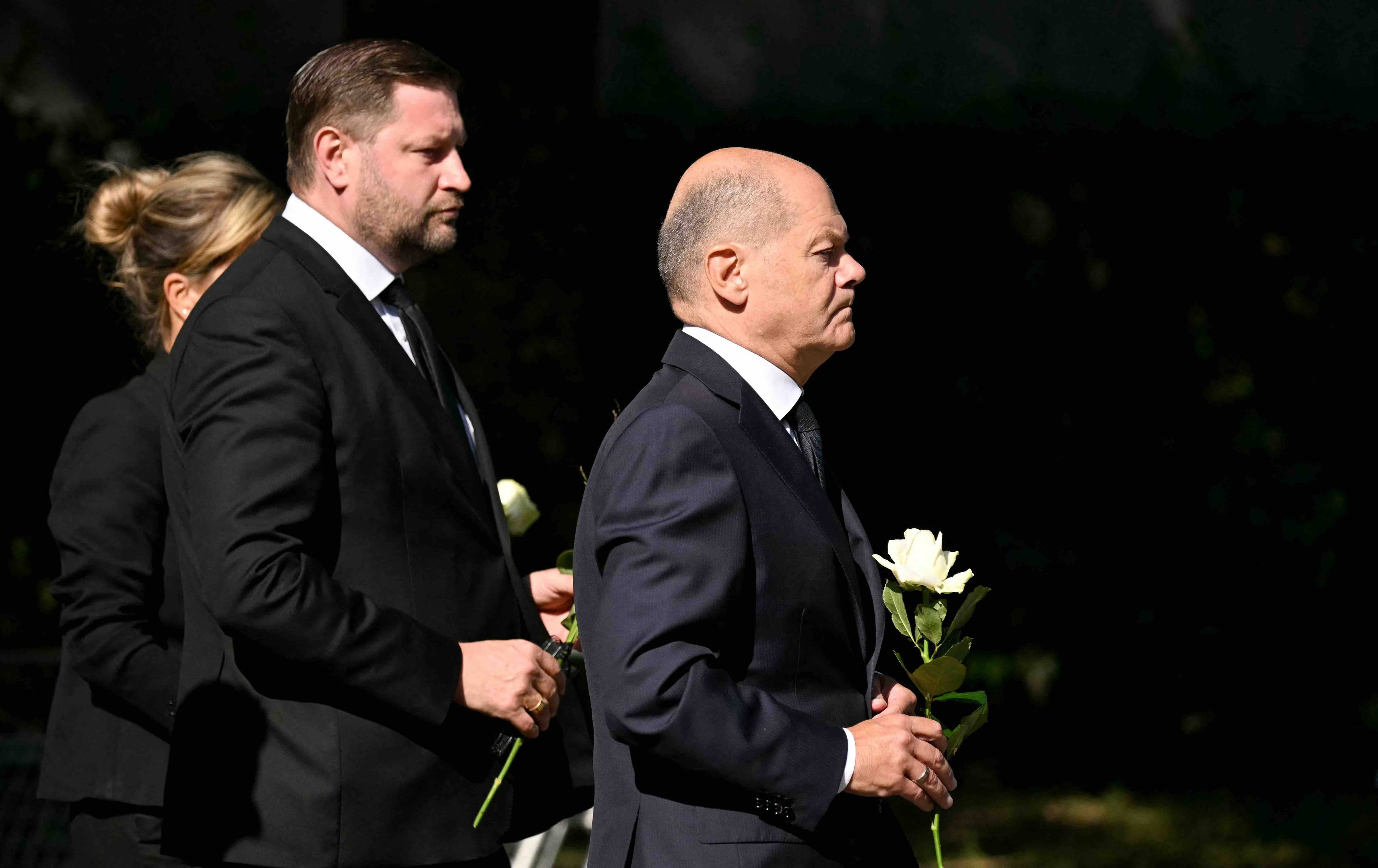 Olaf Scholz stoi trzymając w ręku biały kwiat, za nim stoi mężczyzna w ciemnym garniturze, także z kwiatem. Solingen