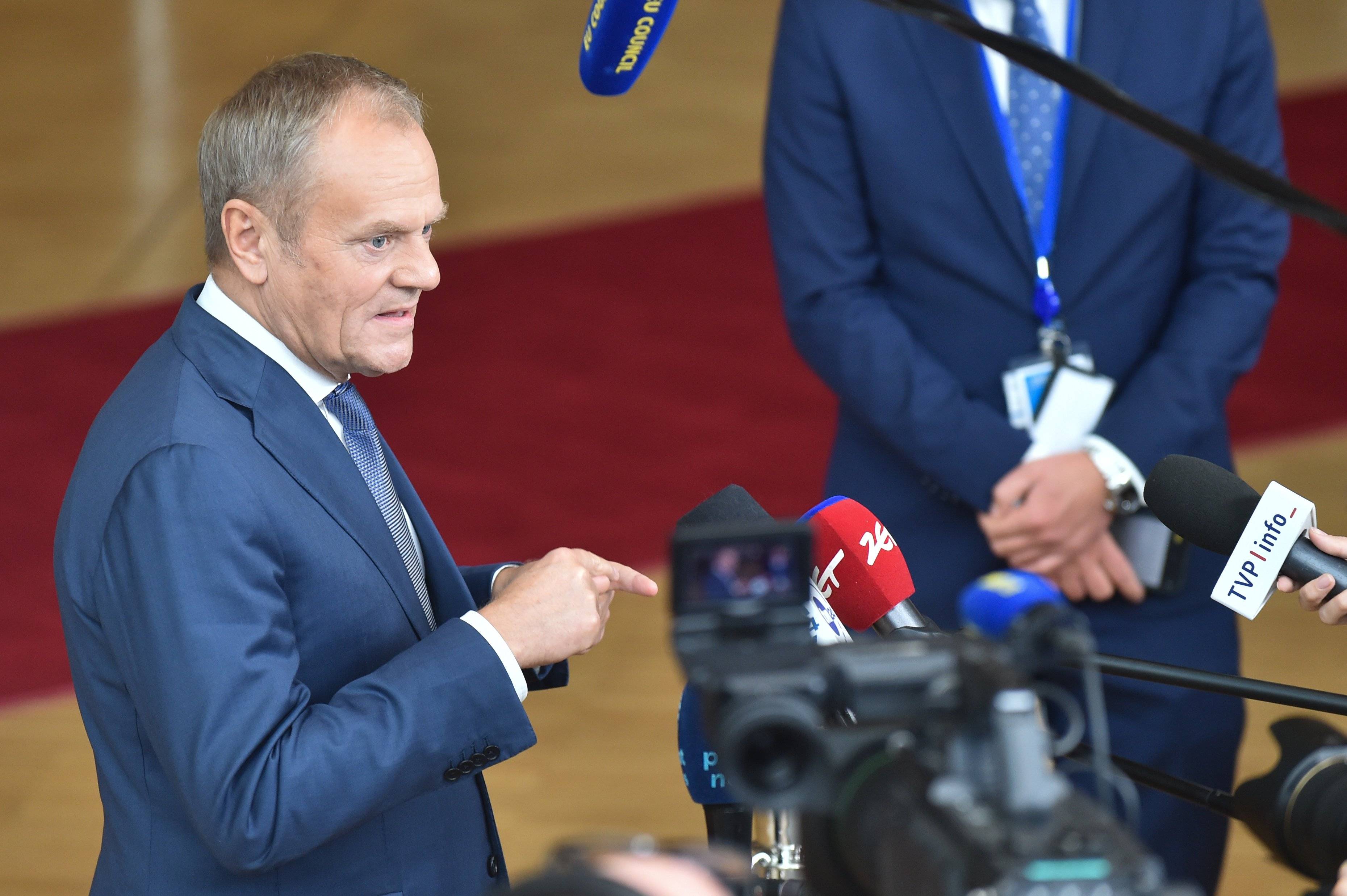 Premier Donald Tusk w niebieskim garniturze stoi przed mikrofonami dziennikarzy w holu prowadzącym do sali plenarnej budynku Rady Europejskiej, w tle widać czerwony dywan.