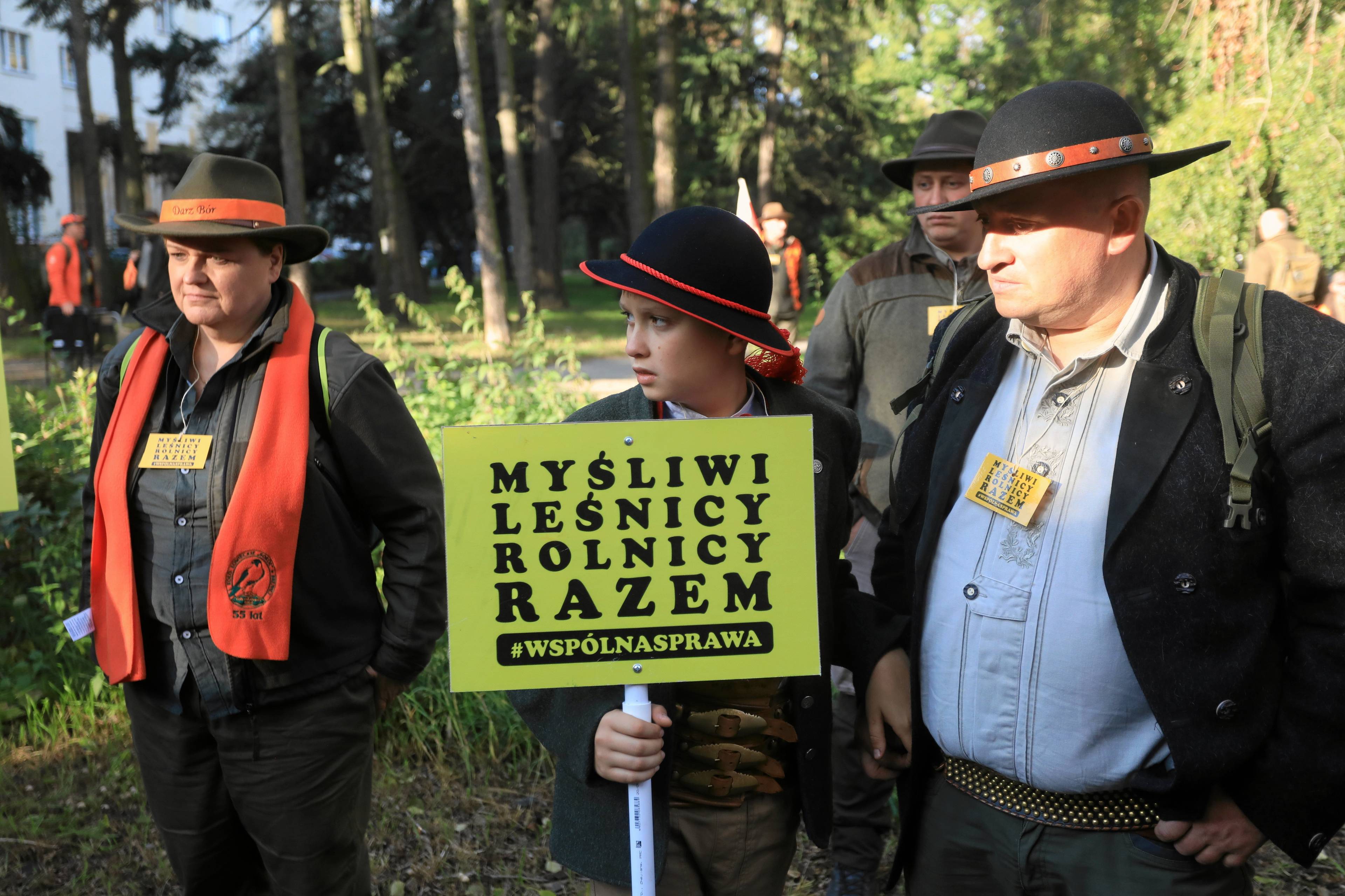 Dwaj męzczyźni i chlopiec w kapeluszach trzumają transparent "Myśliwi, rolnicy razem. Wspólna sprawa"