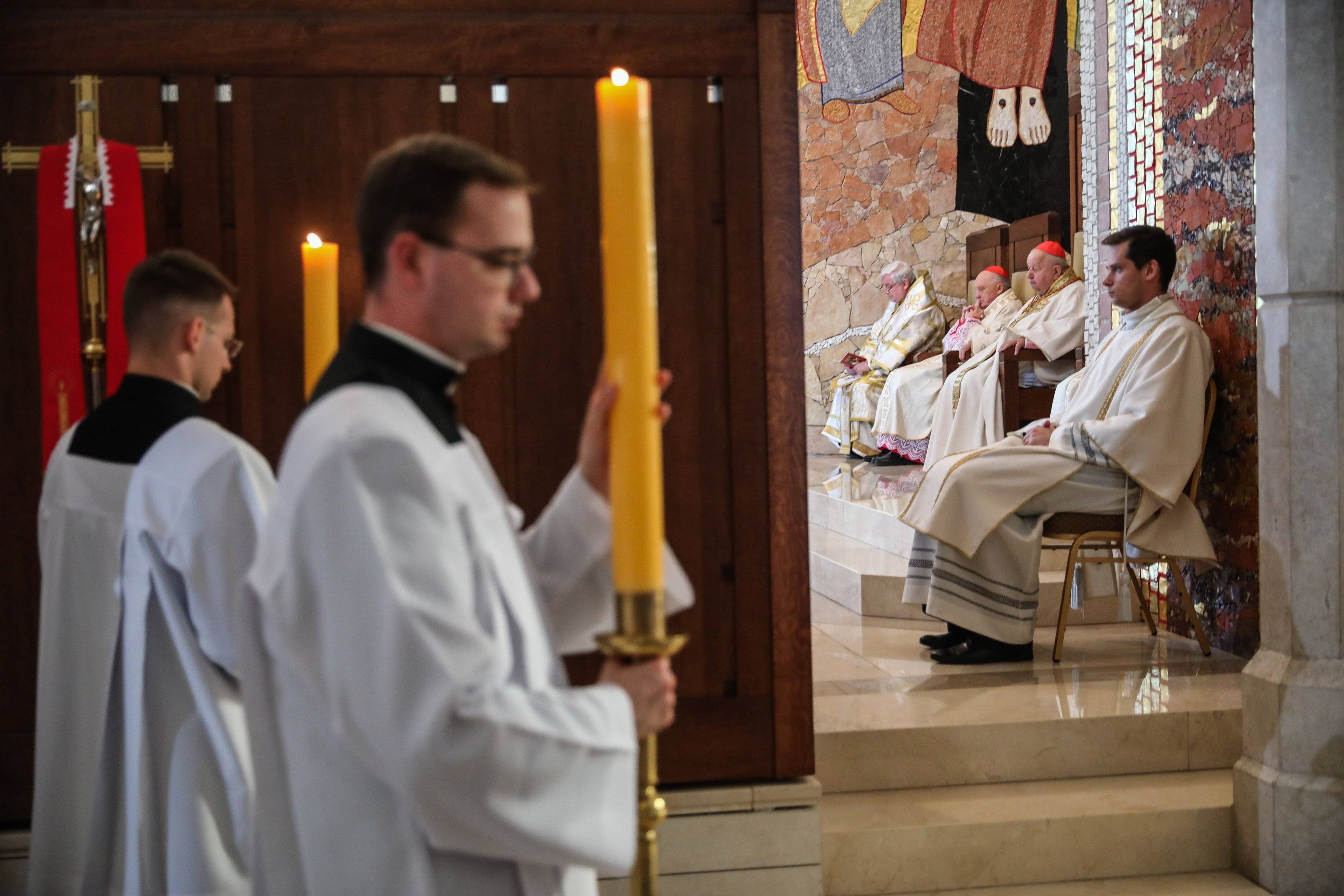 Dostojnicy kościelni siedzą w fotelach w czasie uroczystosci religijnej, a księża wnoszą wysokie świece