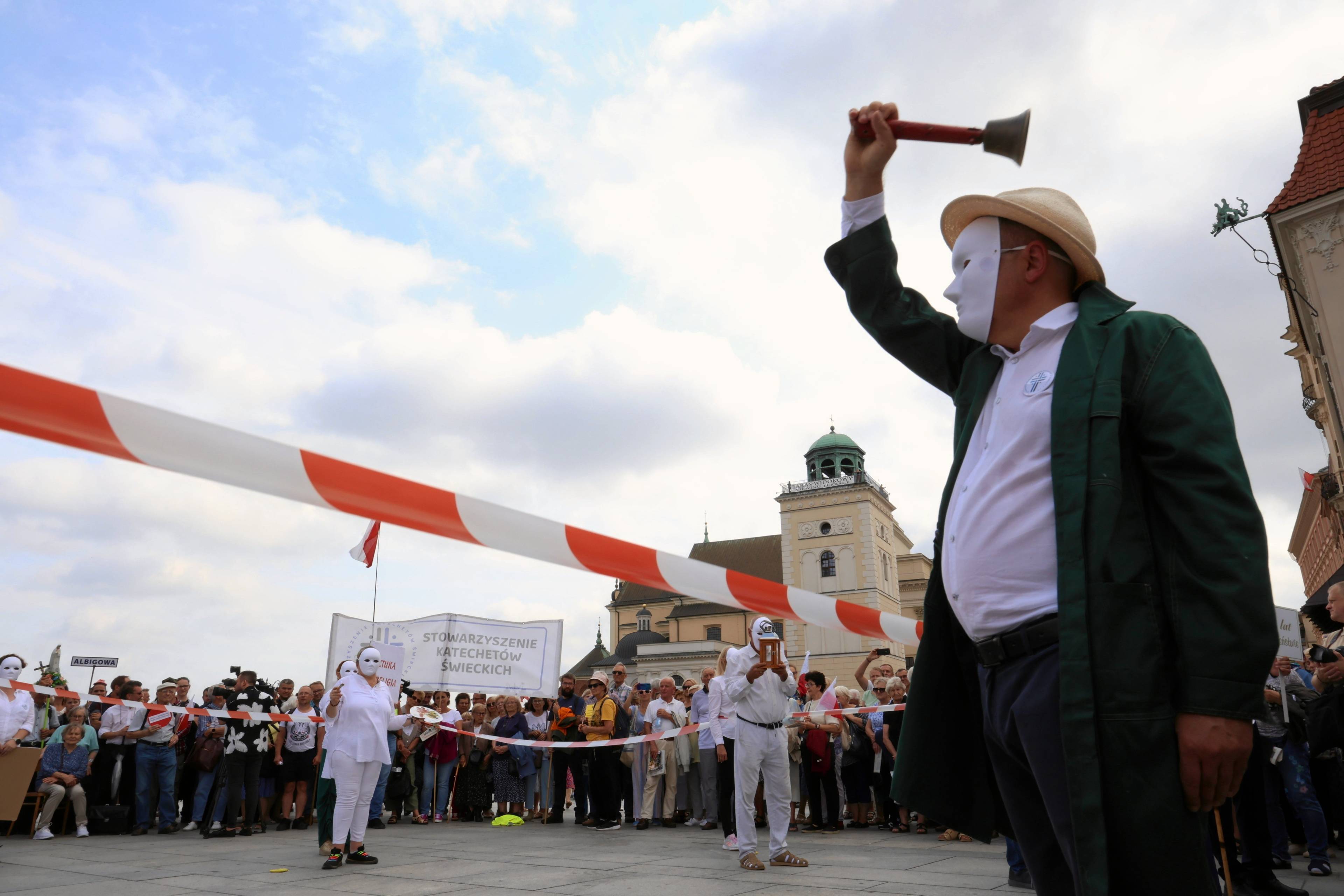 Mężczyzna w białek masce zakrywającej całą twarz macha szkolnym dzwonkiem