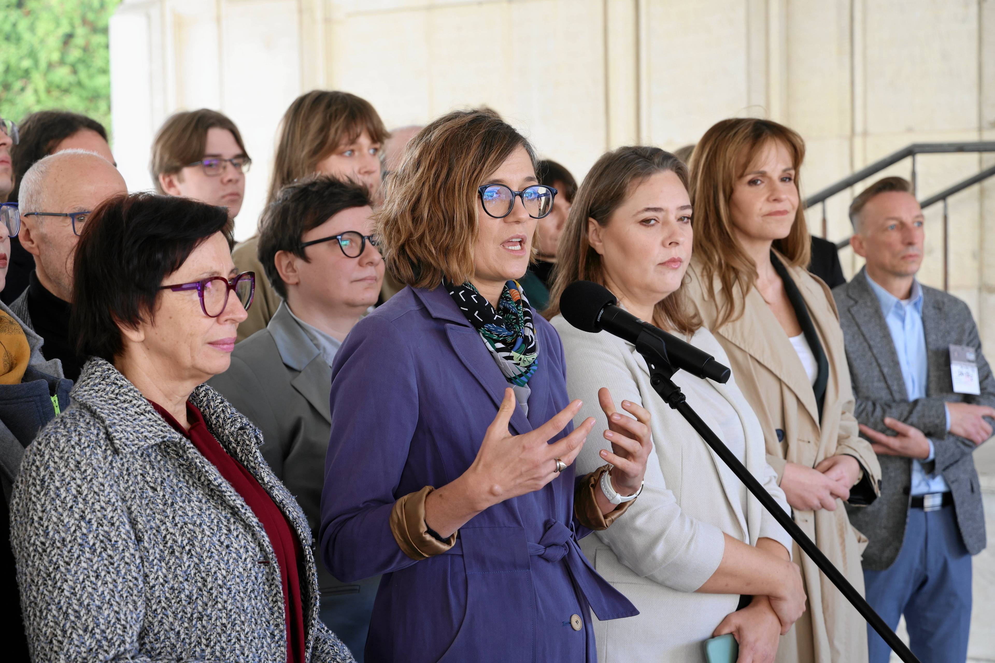 Magdalena Biejat w fioletowym płaszczu przemawia przez mikrofon przed budynkiem Senatu. Obok niej stoi senatorka Anna Górska. Na zdjęciu widzimy też posłanki - Dorota Olko, Joanna Wicha. Na drugim planie działacze i działaczki Lewicy Razem