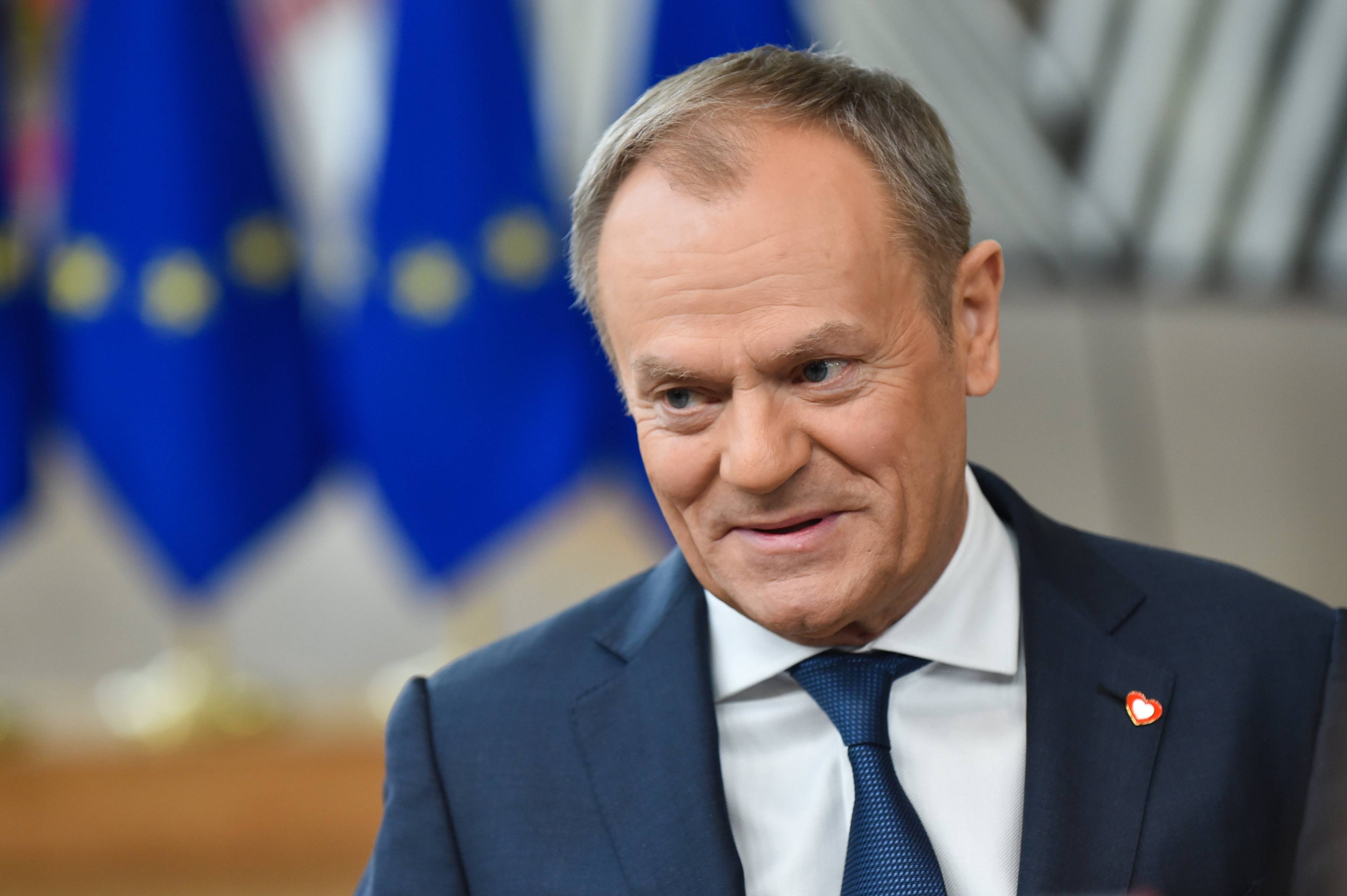 Premier Polski Donald Tusk w niebieskim garniturze i niebieskim krawacie uśmiecha się rozmawiając z dziennikarzami tuż przed rozpoczęciem szczytu Unii Europejskiej w lutym 2024; w tle niebieska flaga UE z żółtymi gwiazdkami