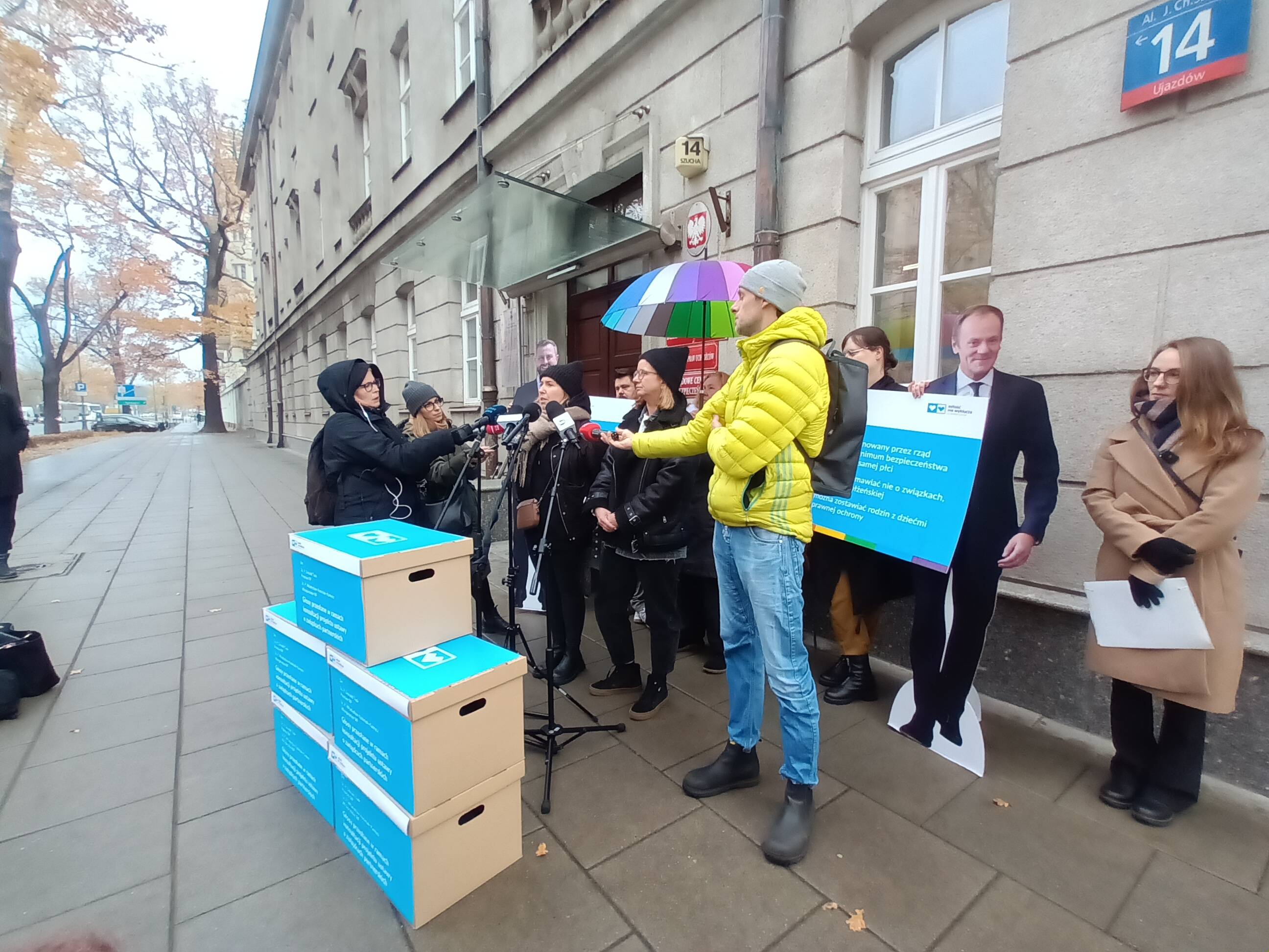 Dwie kobiety przed dziennikarzami i wielkimi pudłami. W tle - wizerunek premiera Tuska naturalnej wielkości