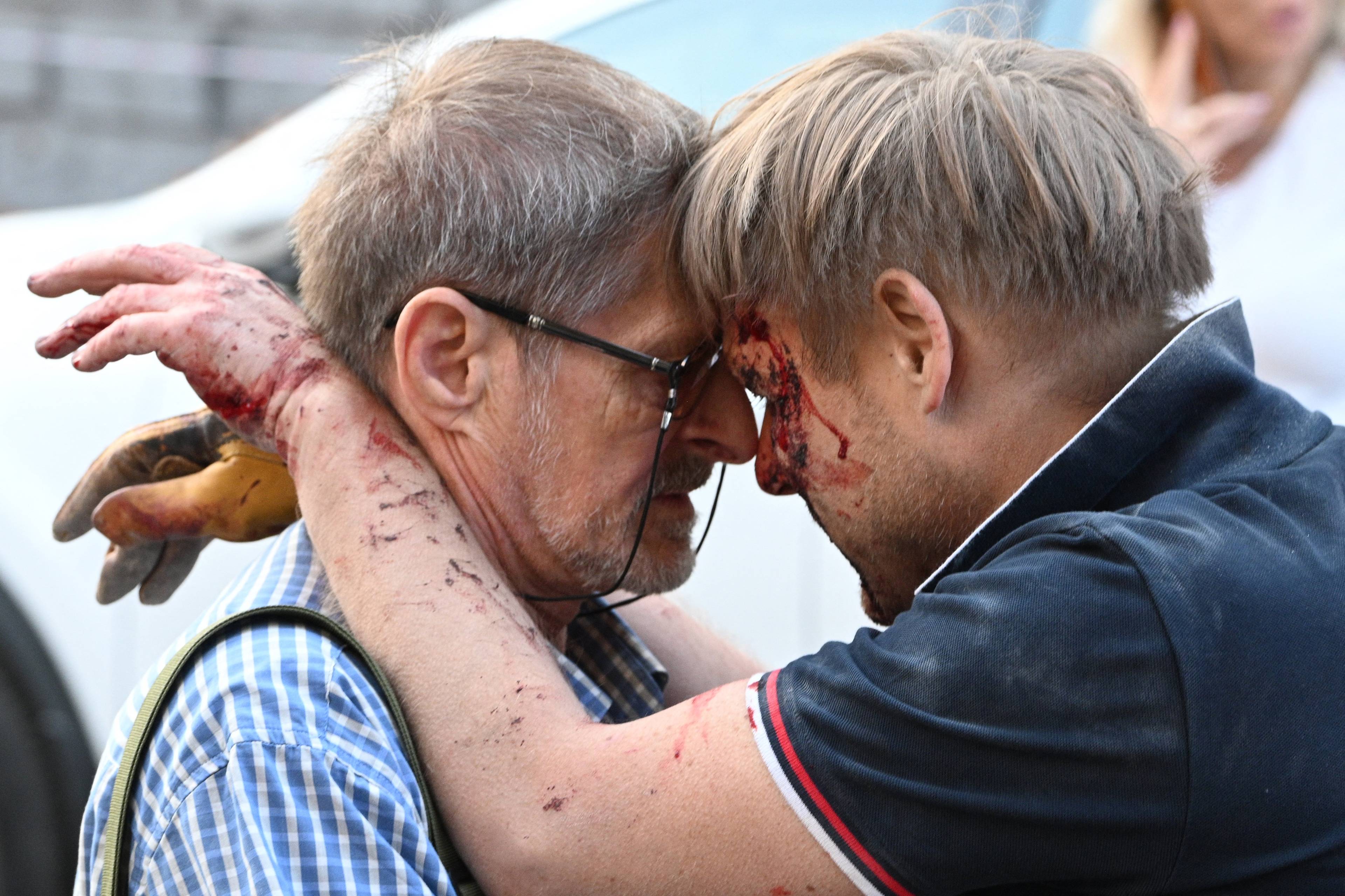 Na zdjęciu widać dwóch mężczyzn. Mężczyzna po prawej opiera się o starszego mężczyznę po lwej. Jest w czarnej koszuli, ma zakrwawioną twarz i ręce.