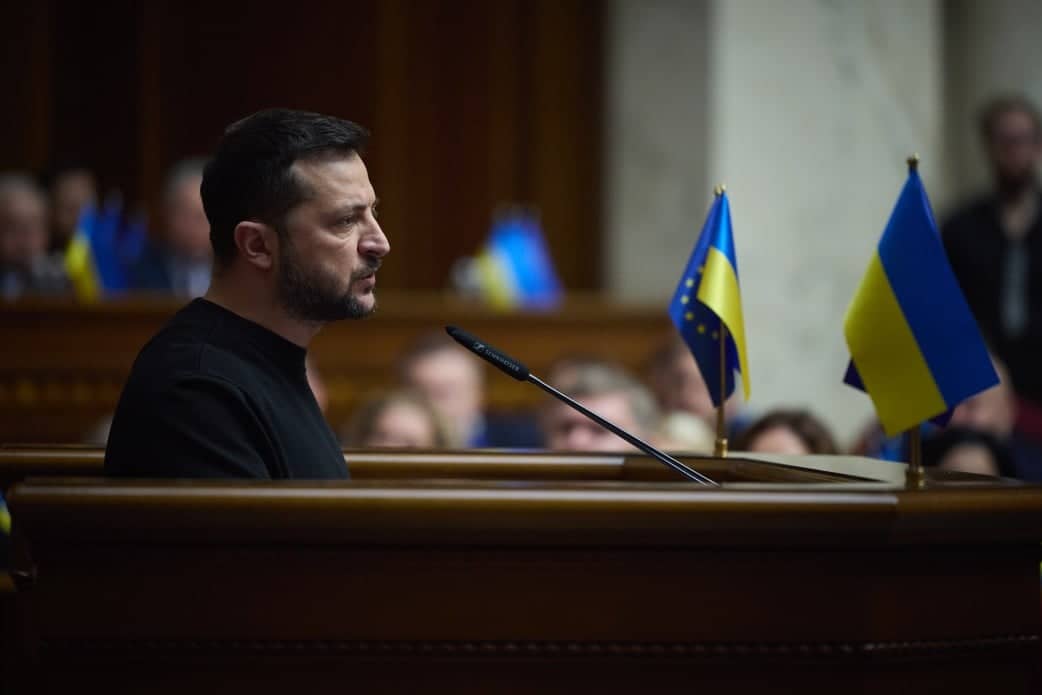 Na zdjęciu jest mężczyzna ok. 45 lat. Ma czarne krótkie włosy i brodę. Jest ubrany w czarną bluzę. Stoi przy ambonie. Po prawej stronie na ambonie są małe flagi- ukraińska oraz Unii Europejskiej