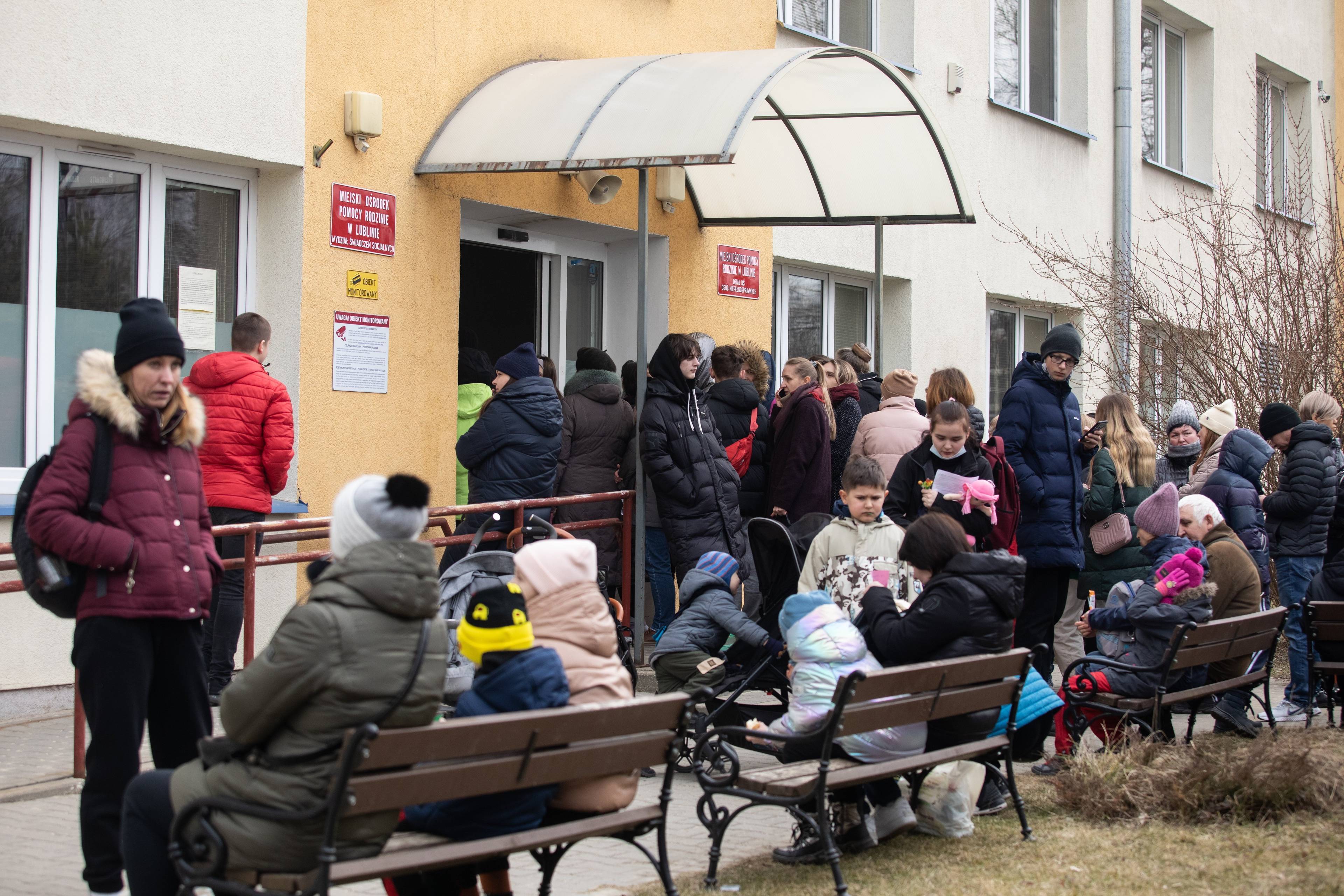 Na zdjęciu widać ludzi, którzy stoją w kolejce obok pomarańczowego budynku, niektórzy siedzą na ławkach obok. Wśród nich są dzieci