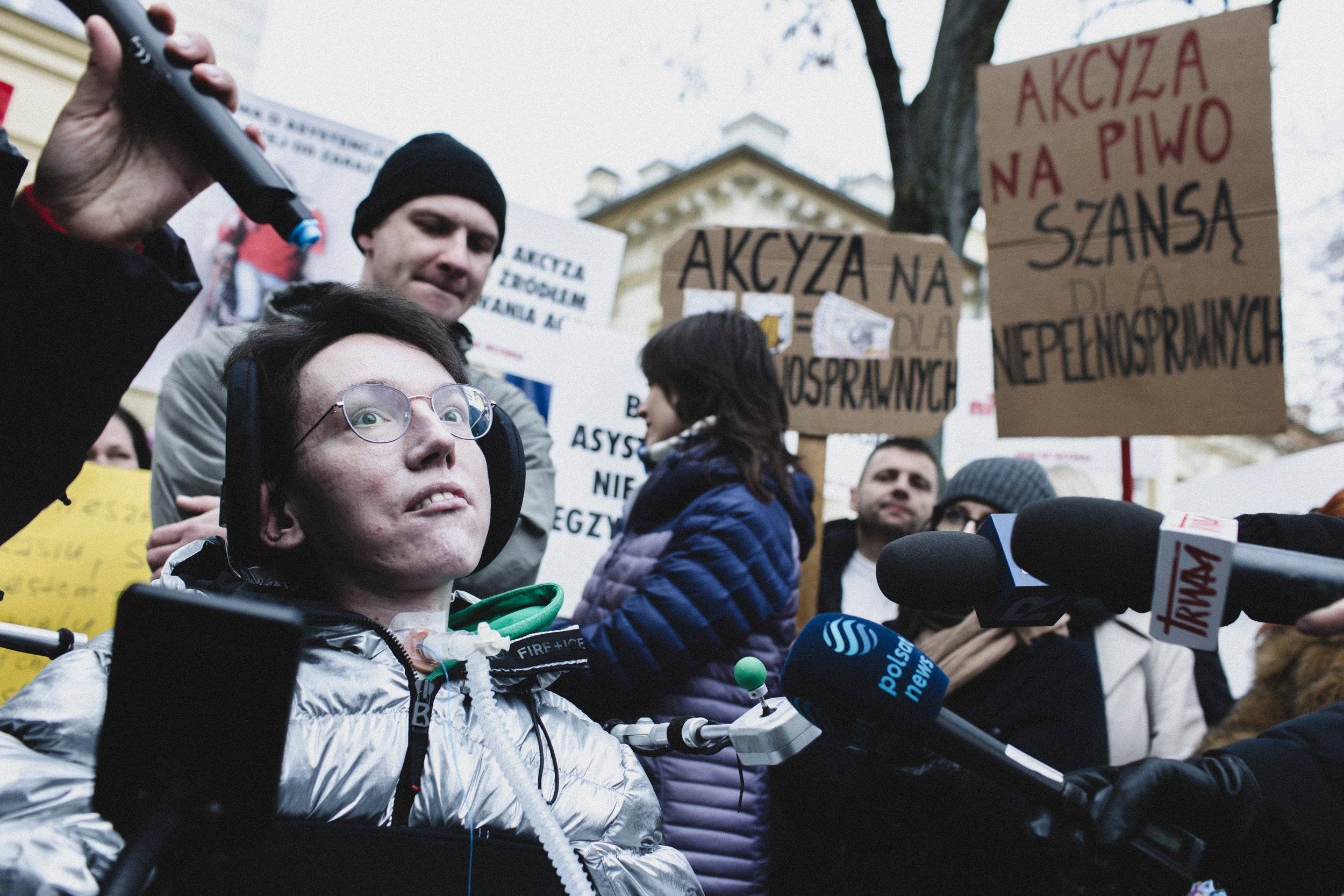 03.12.2024, Warszawa. Chorujący na dystrofię mięśniową inluencer Wojtek Sawicki na proteście osób z niepełnosprawnościami pod KPRM. OzN domagają się ustawy o asystencji osobistej. Na zdjęciu siedzący na wózku inwalidzkim młody mężczyzna w okularach z podłączoną do krtani rurką do tracheotomii. Za nim hasło na kartonowym transparencie "Akcyza na piwo szansą dla niepełnosprawnych”