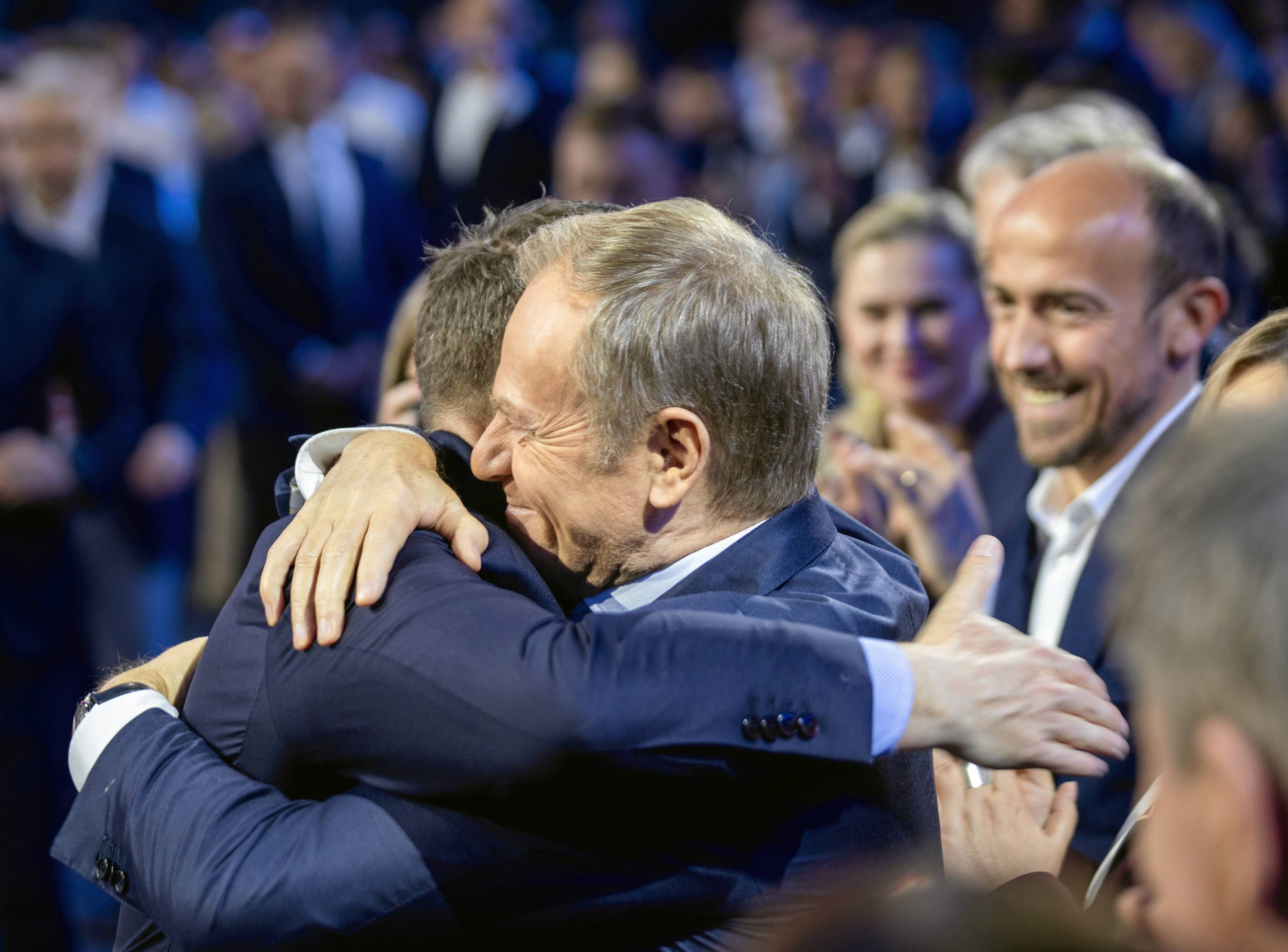 Dwóch mężczyzn w uścisku, za nimi uśmiechnięci ludzie