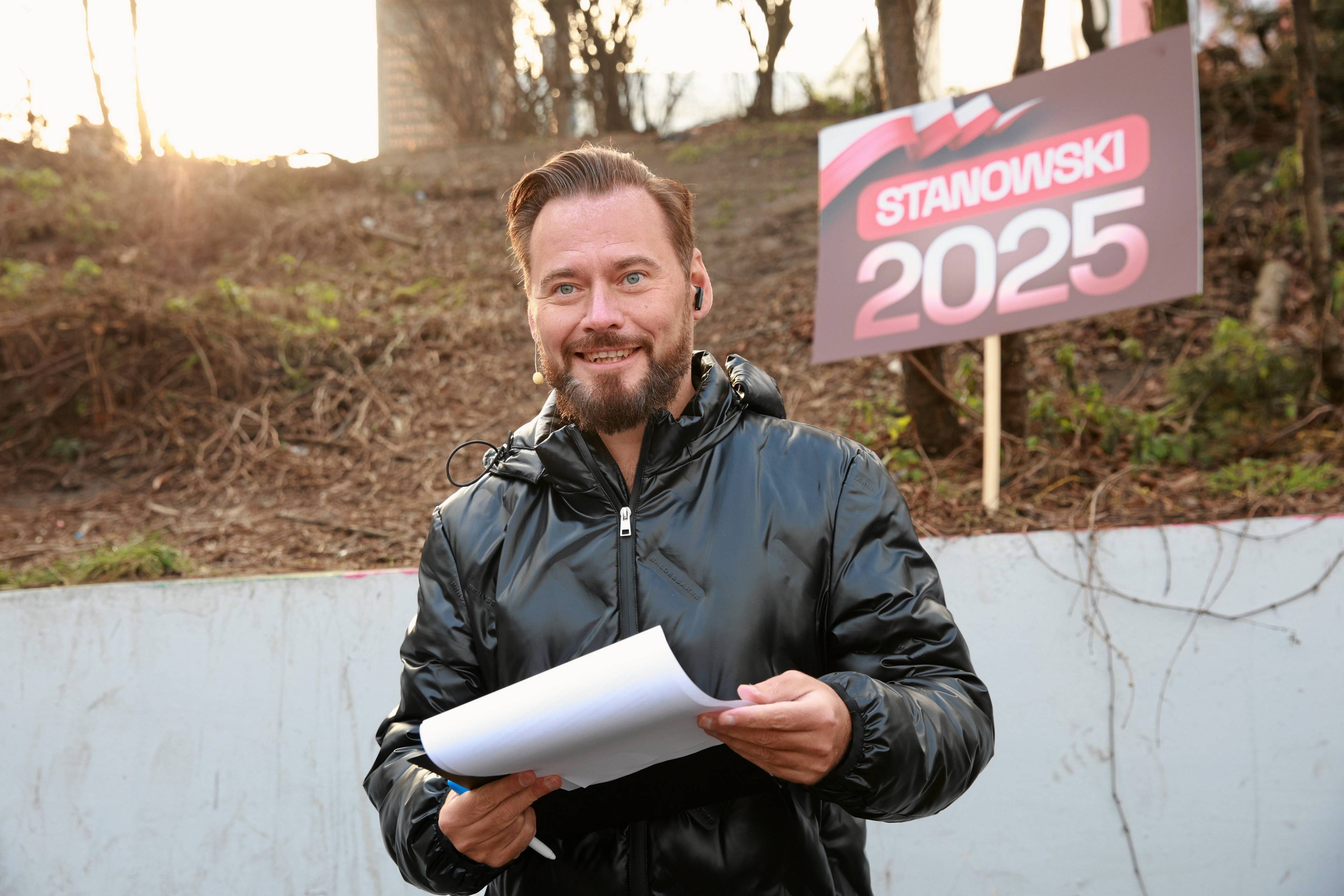 Uśmiechnięty mężczyzna z kartką i banerem z napisem „2025” za plecami. Krzysztof Stanowski