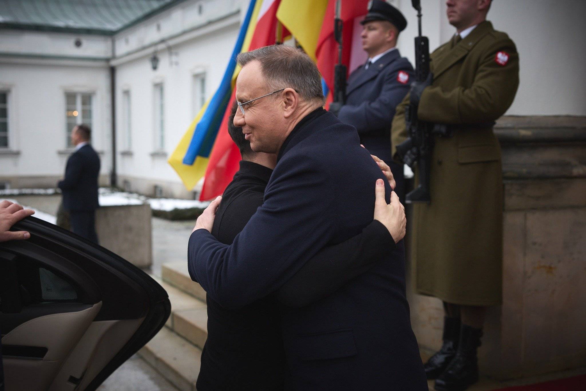 na zdjęciu jest czterech mężczyzn. Mężczyzna w czarnym płaszczu ściska mężczyznę w czarnej bluzie. Z tyłu stoi dwóch żołnierzy w mundurach polskiej armii