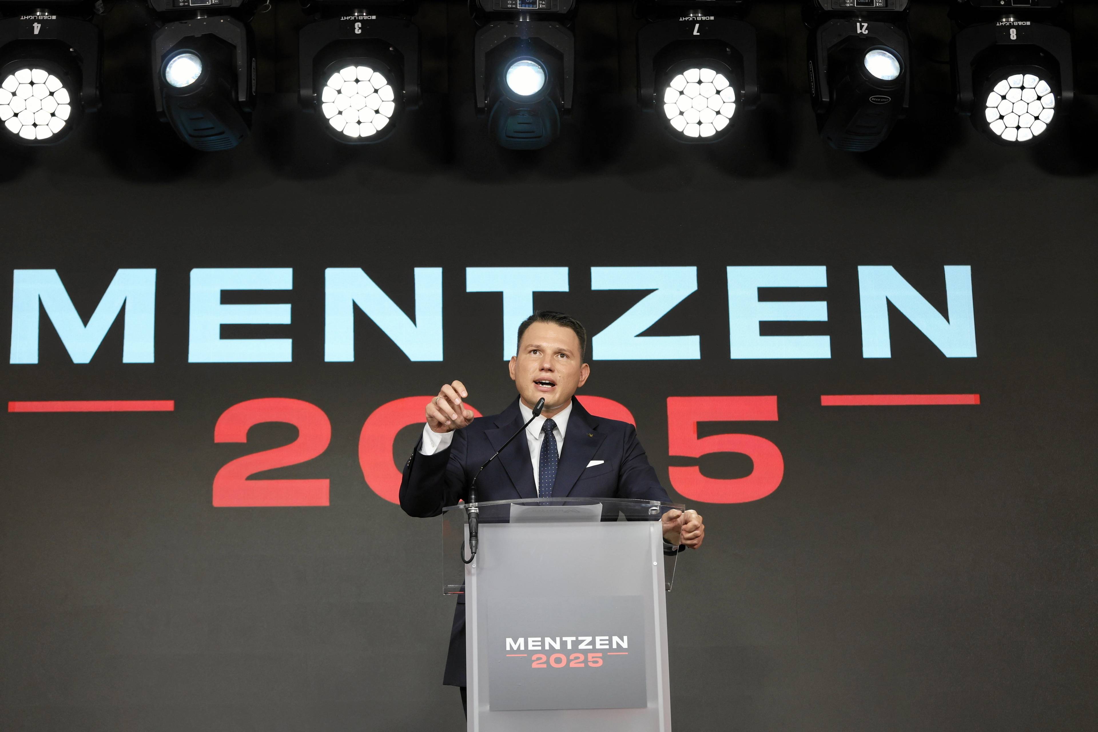 Mężczyzna w garniturze i białej koszuli stoi za mównicą i przemawia, na tle dużego napisu "Mentzen 2025"