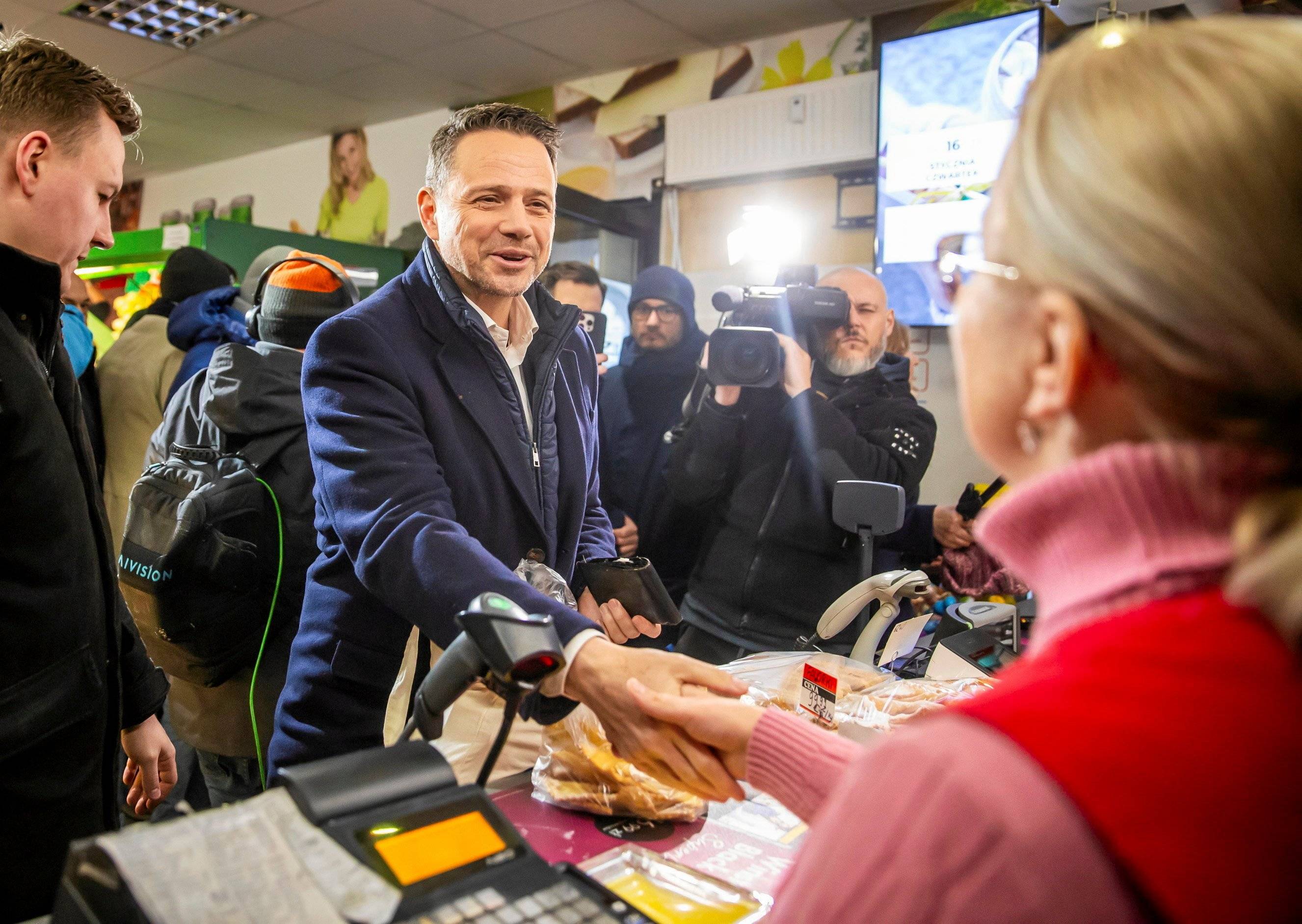 Mężczyzna uśmiecha się do kasjerki w sklepie, za nim mężczyzna z kamerą