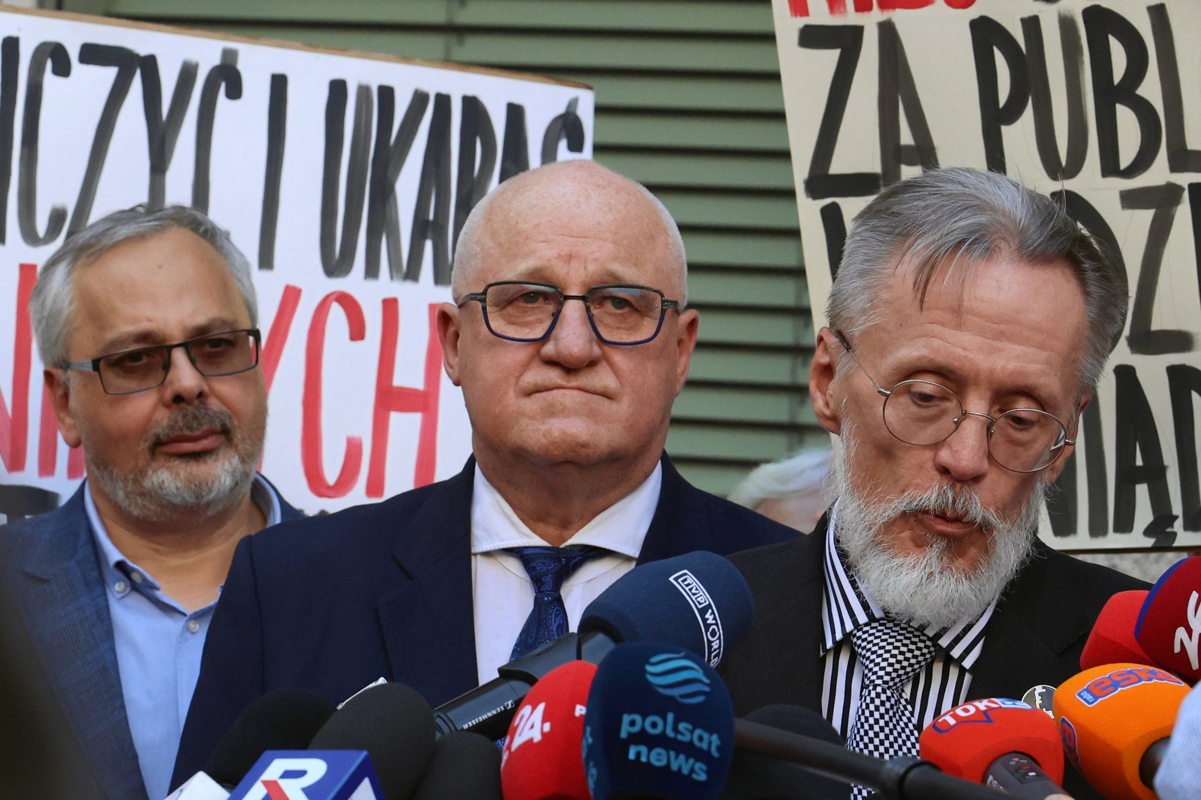 Trzech mężczyzn przed mikrofonem, w tle protest