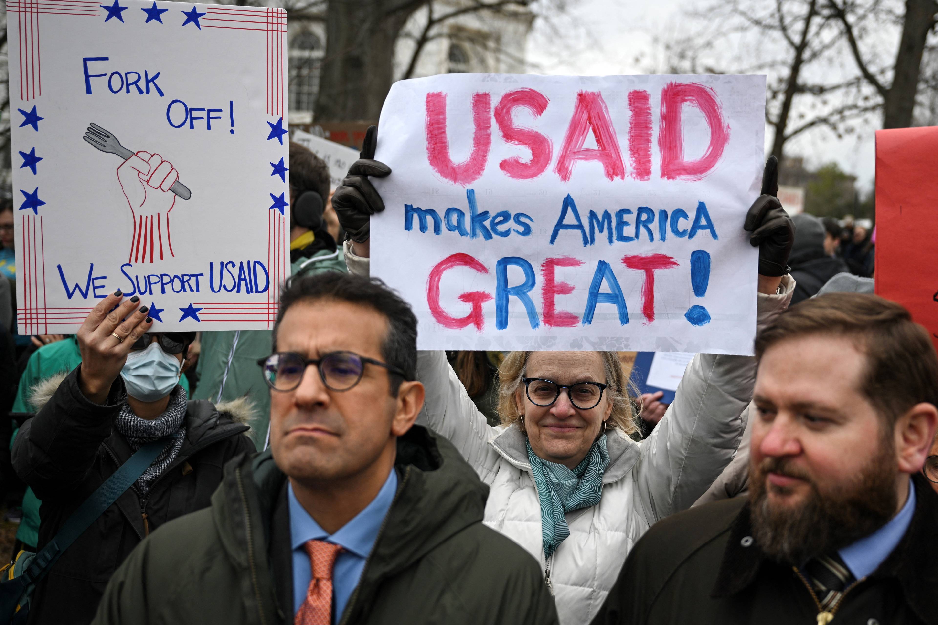 Tłum ludzi na demonstracji, jedna z kobiet trzyma nad głową biały transparent z napisem "USAID makes America GREAT!"