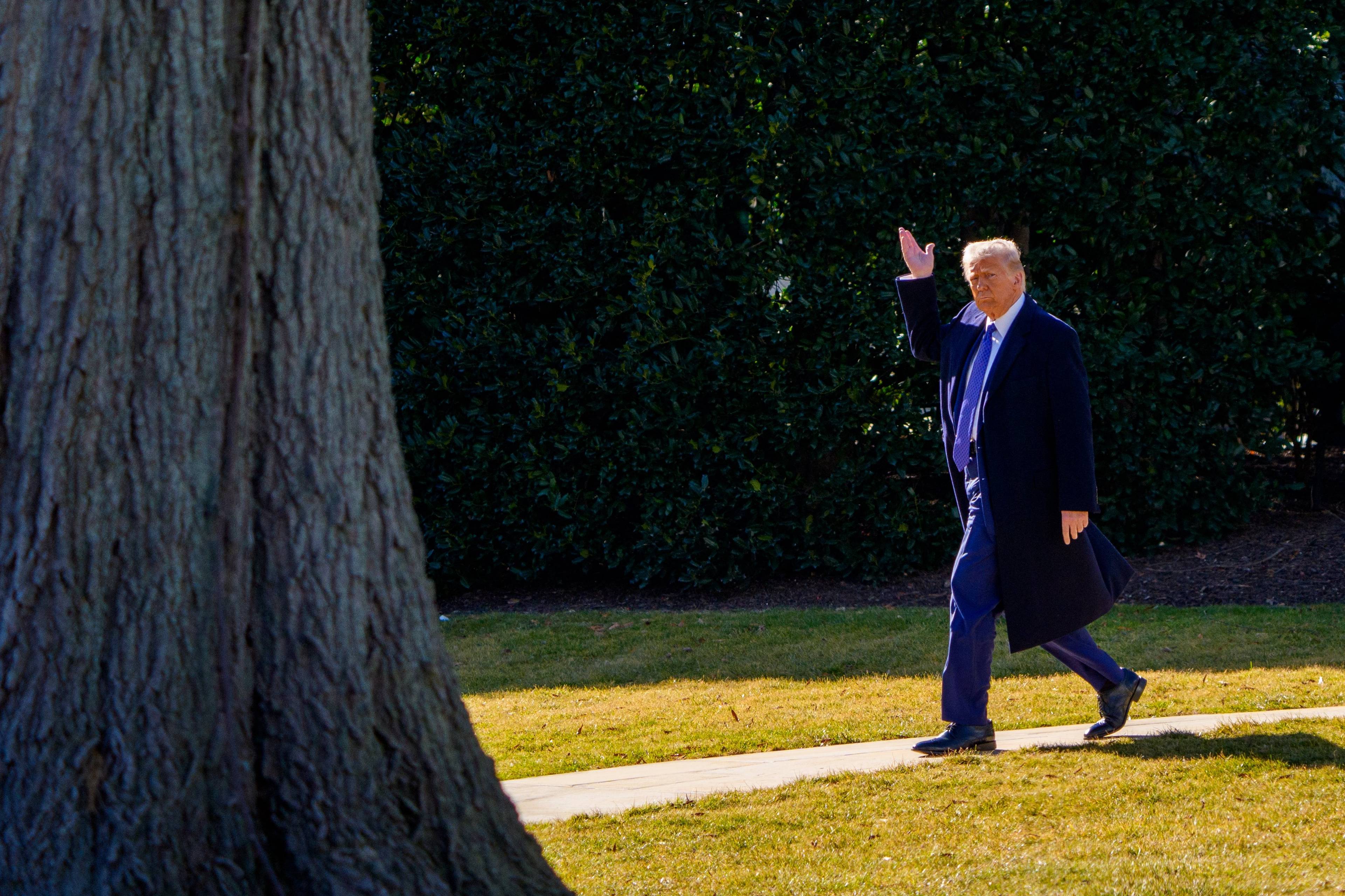Donald Trump idzie chodnikiem na tle zieleni, jest ubrany w płaszcz, macha w stronę fotoreportera