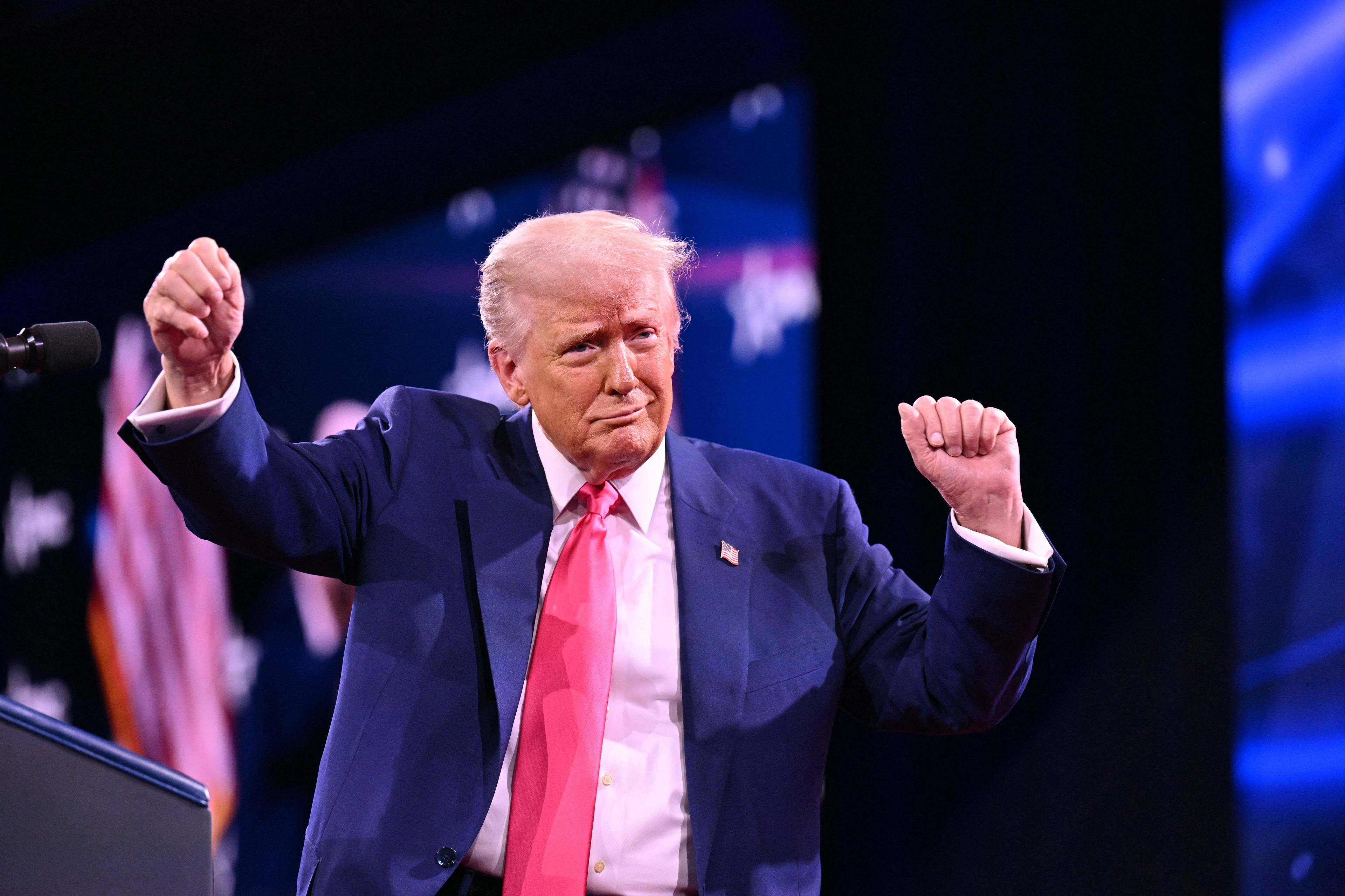 Starszy mężczyzna w niebieskim garniturze i różowym krawacie (prezydent USA Donald Trump) tańczy na scenie z uniesionymi w górę rękami