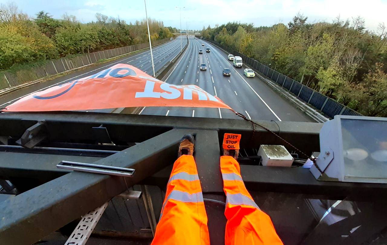 Na zdjęciu widać nogi w pomarańczowym, odblaskowym kombinezonie stojące na ramie nad autostradą. Do podeszwy buta przymocowany jest pomarańczowy napis: "Just Stop Oil"