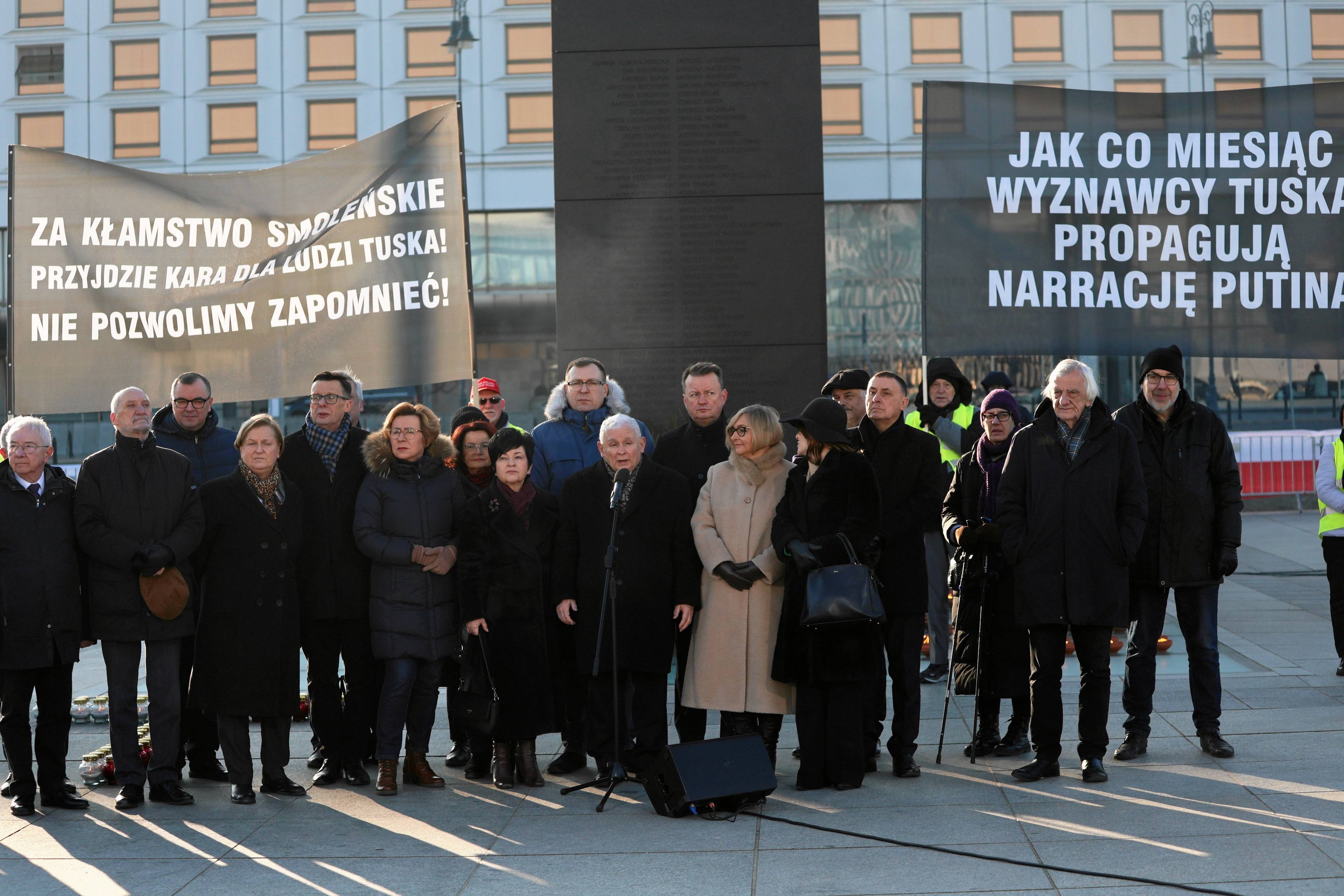 Grupa ludzi w zimowych okryciach stoi na placu, pośrodku niski mężczyzna, Jarosław Kaczyński, przemawia