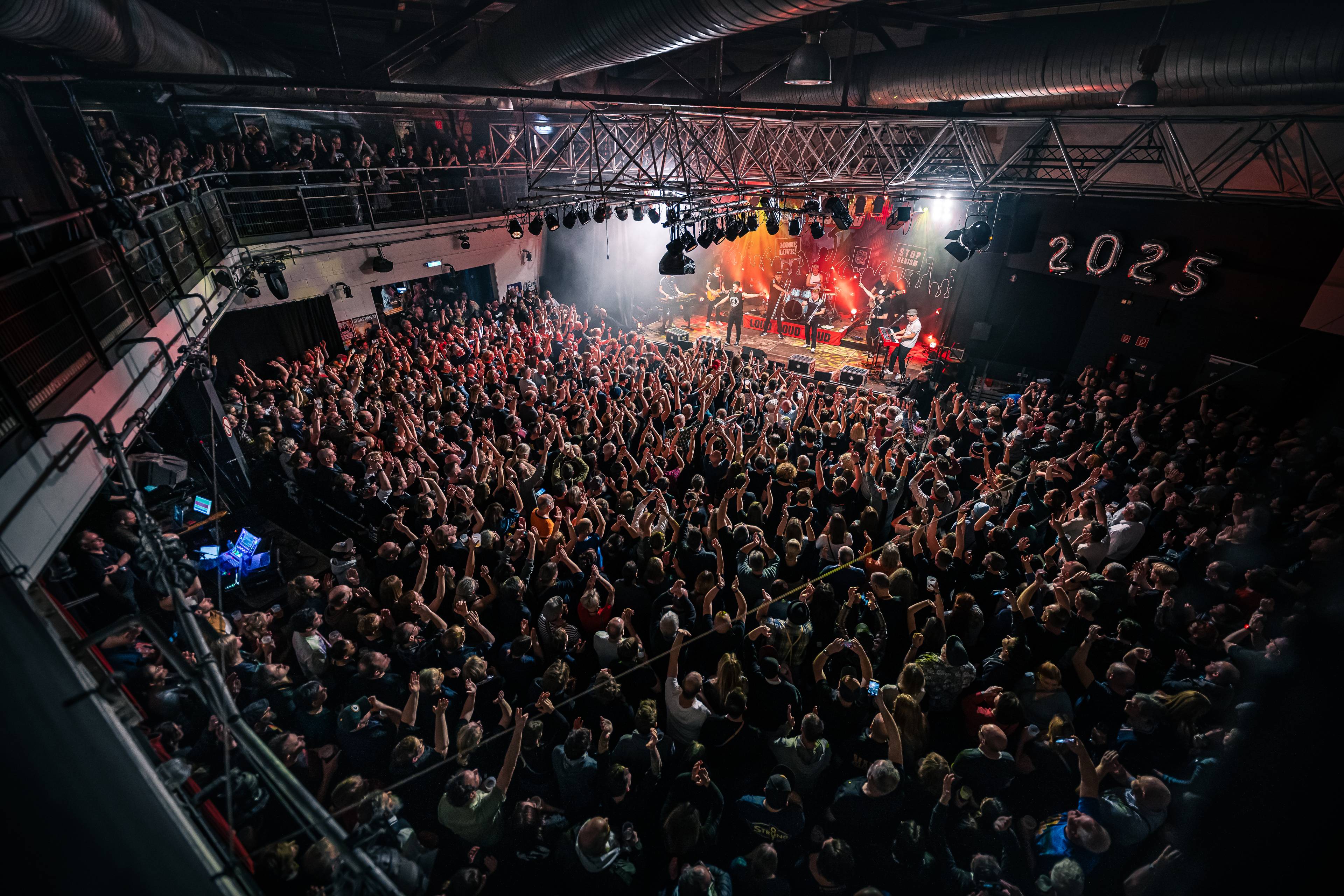 Koncert rockowy w dużej hali. Z boku sceny: "2025"