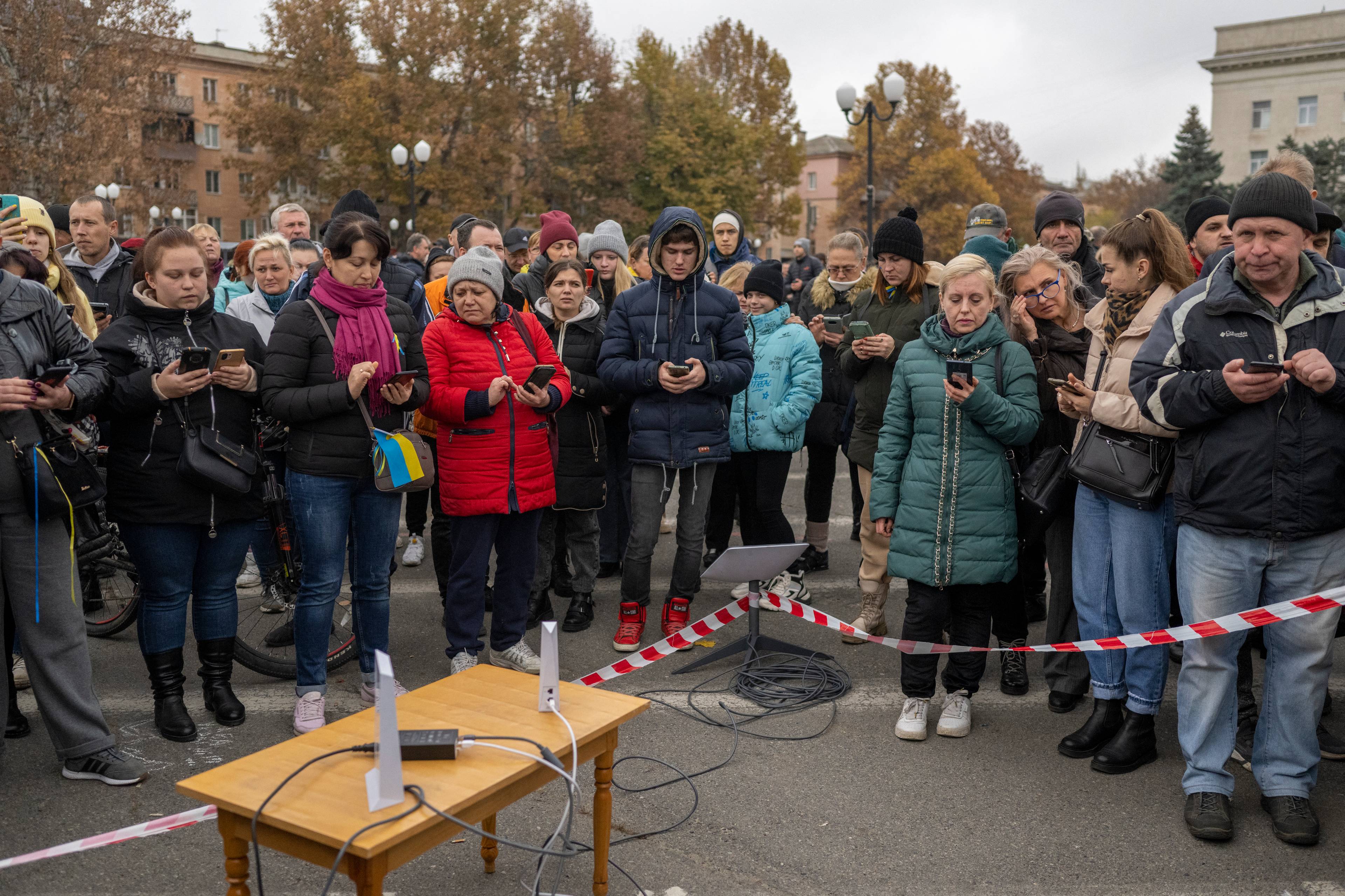 Osoby w zimowych kurtkach stoją wokół stołu z modemami internetowymi. Mieszkańcy Chersonia podłączają telefony do terminala Starlinka