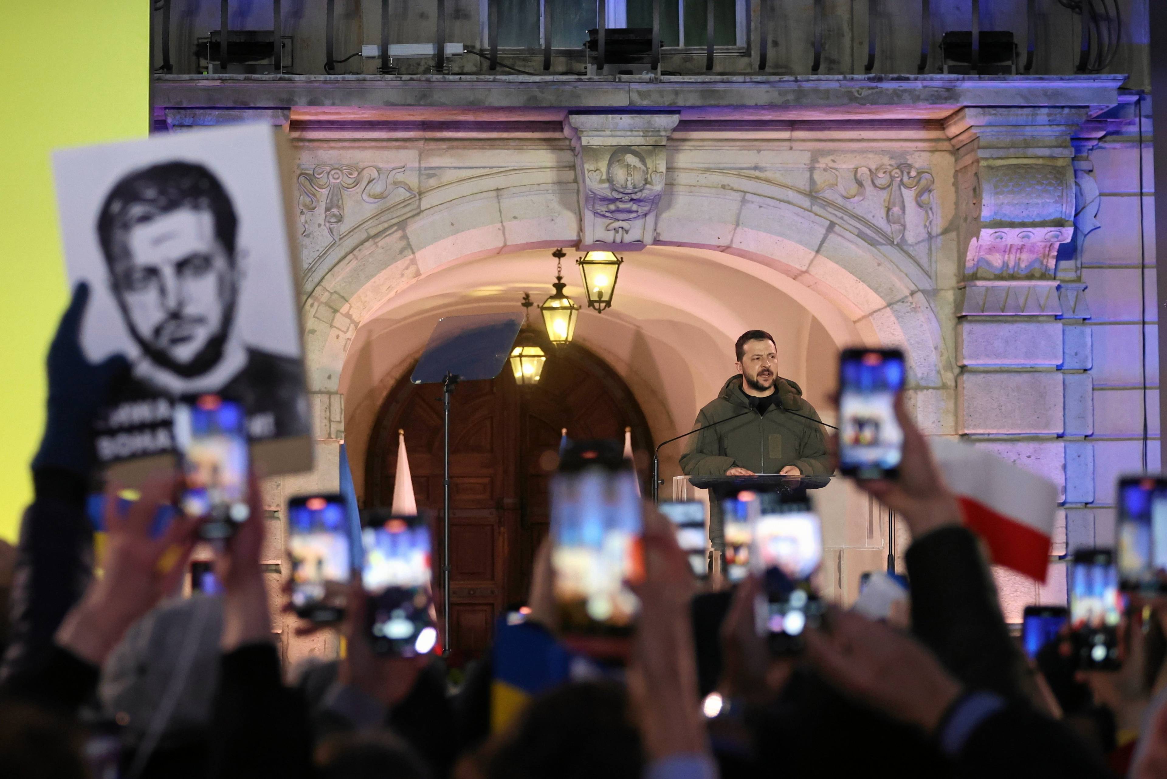 Dziedziniec Zamku Królewskiego, wieczór, oświetlenie, z podwyższenia przemawia mężczyzna, to prezydent Ukrainy Wołodymyr Zełenski