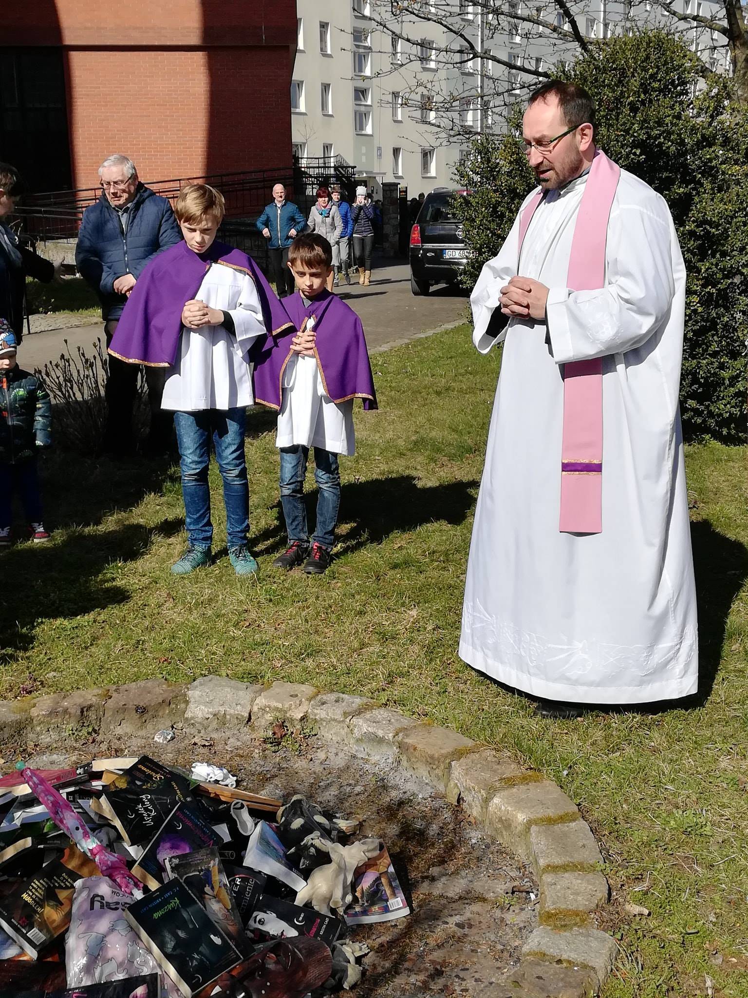 Ksiądz w sztach liturgicznych w różowej stule nad ogniskiem z książkami i maskami