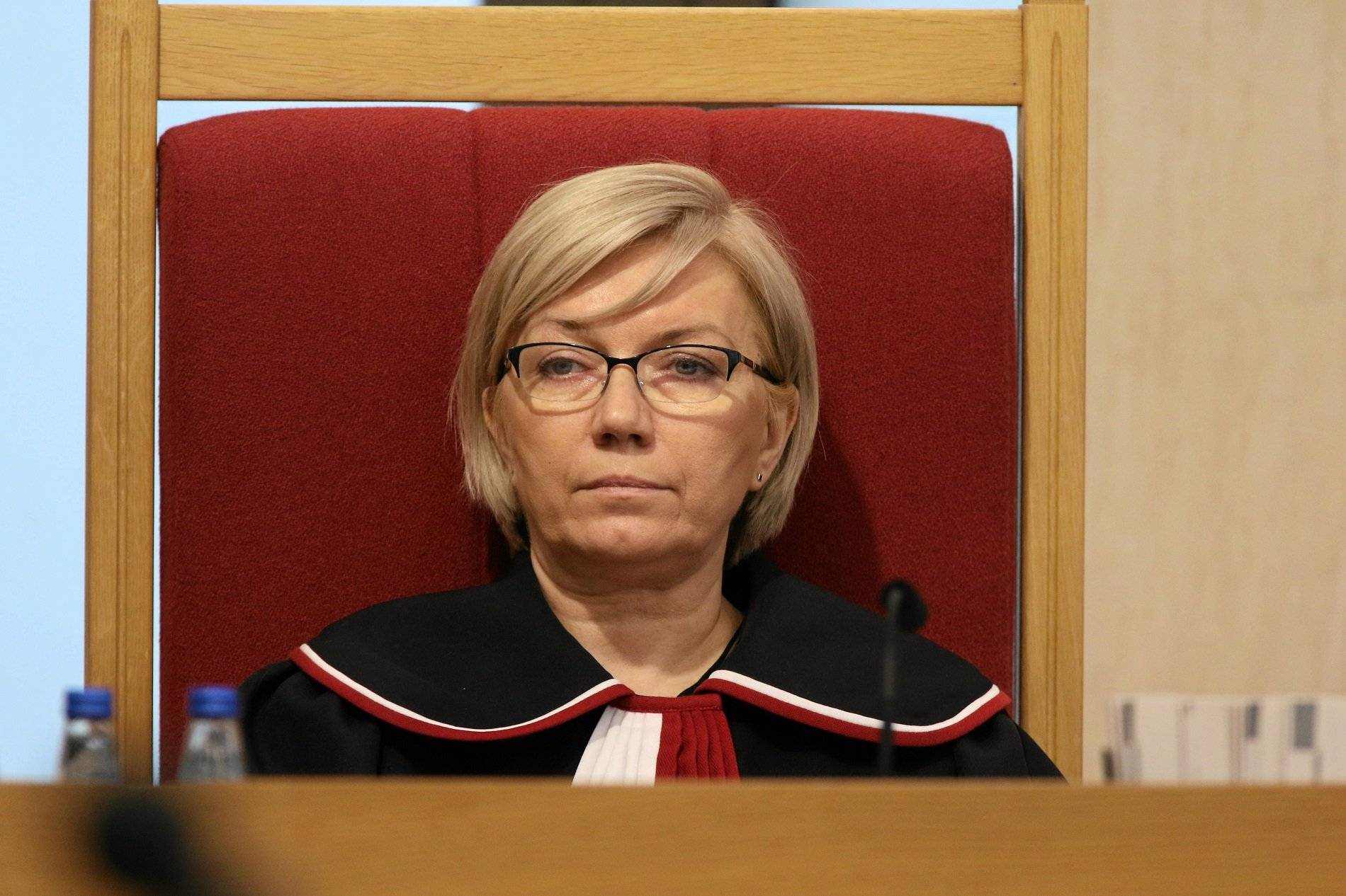 Julia Przyłębska, blondynka z włosami średniej długości, w okularach i w todze sędziowskiej siedzi w czerwonym fotelu