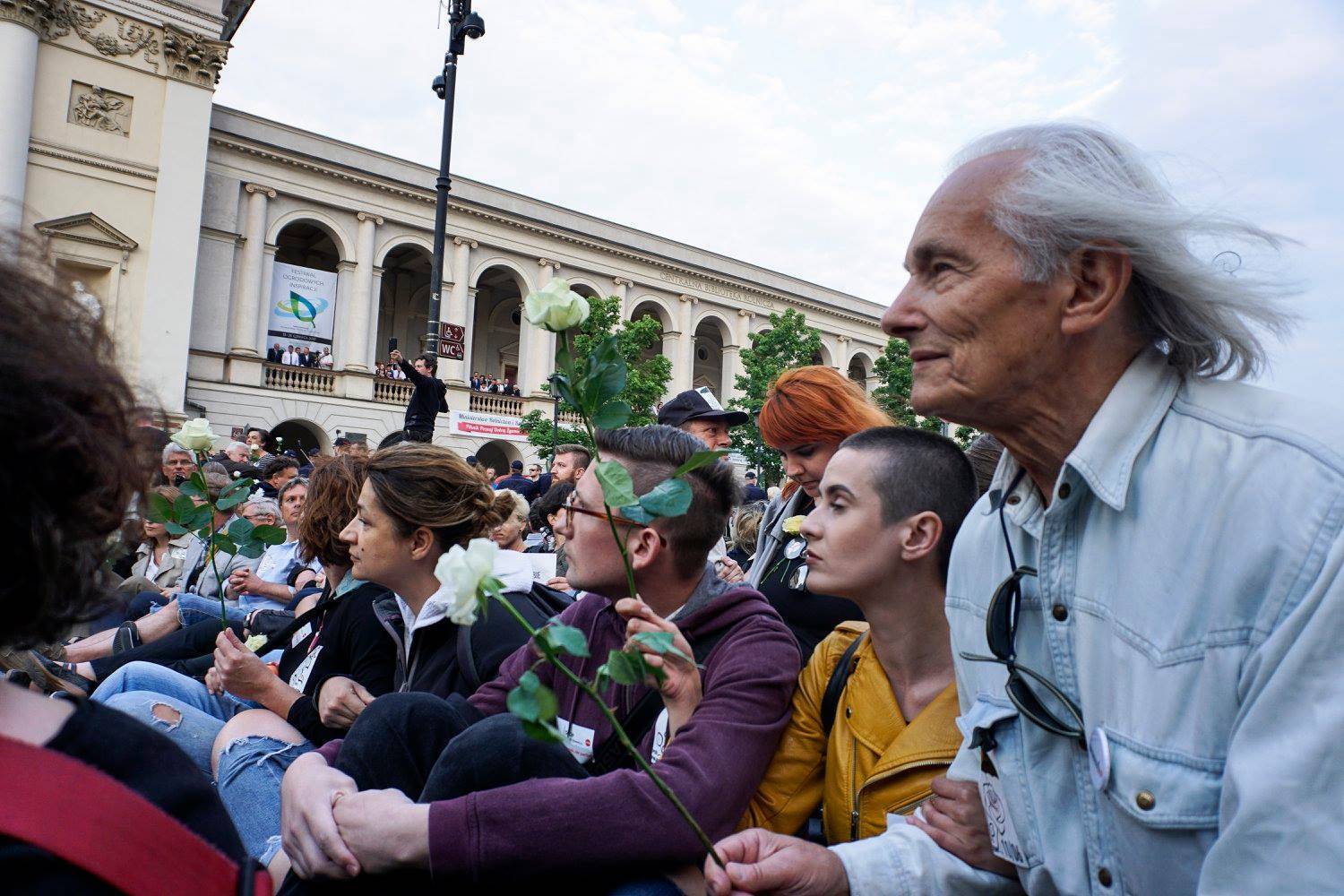 Protestujący siedzą na jezdni Krakowskiego Przedmieścia z białymi różami w rękach