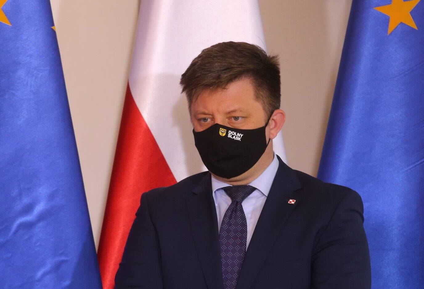 Michał Dworczyk w maseczce na twarzy na tle flag polskiej i unijnej