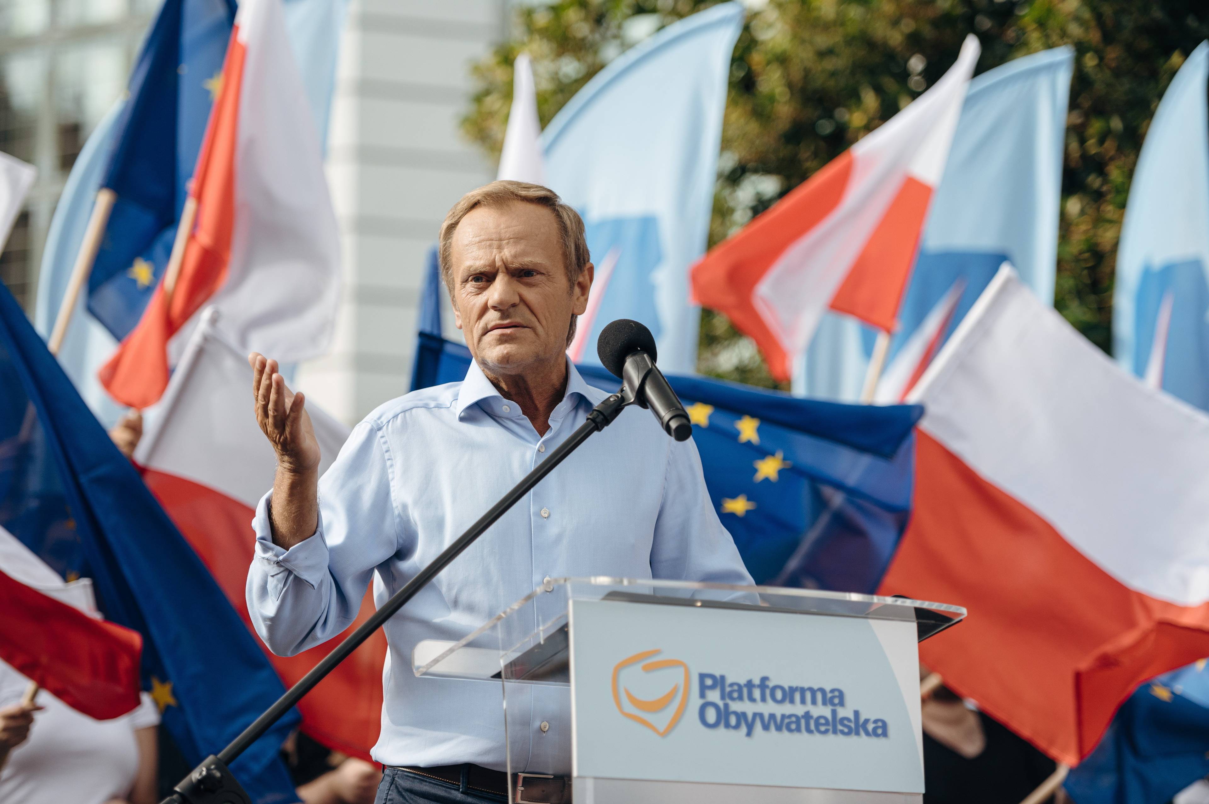 Donald Tusk w jasnej koszuli stoi za mównicą podczas demonstracji. Za nim powiewają flagi Polski i Unii Europejskiej