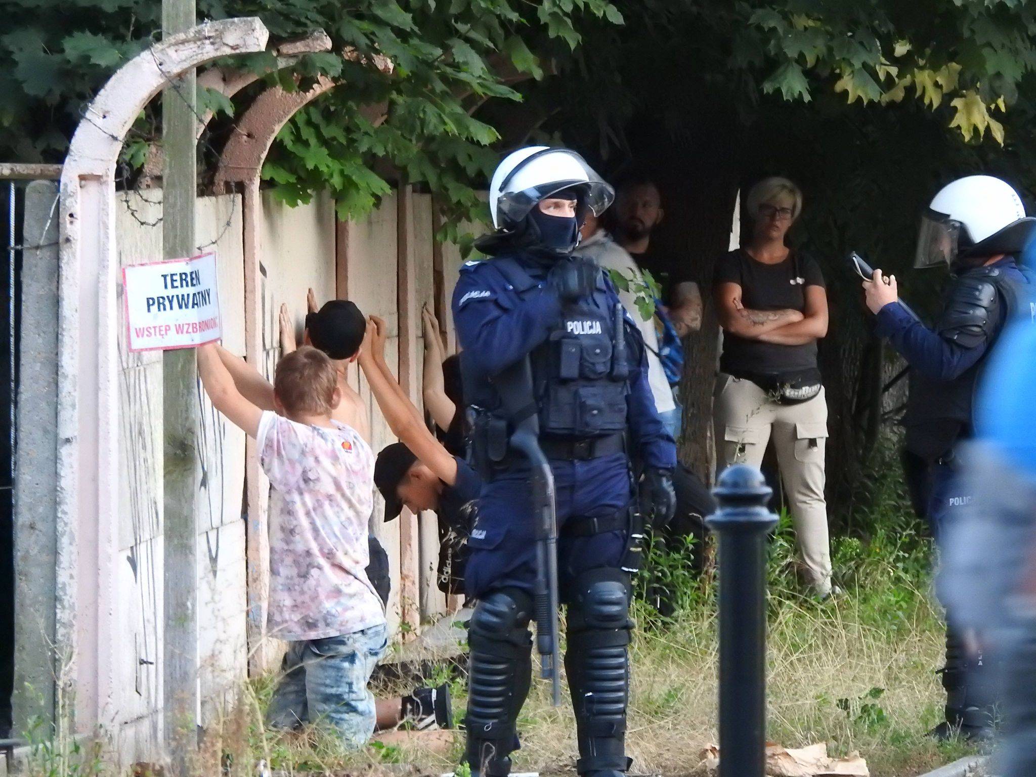 Nastolatki klęczące pod płotem z drutem kolczastym, obok uzbrojeni policjanci