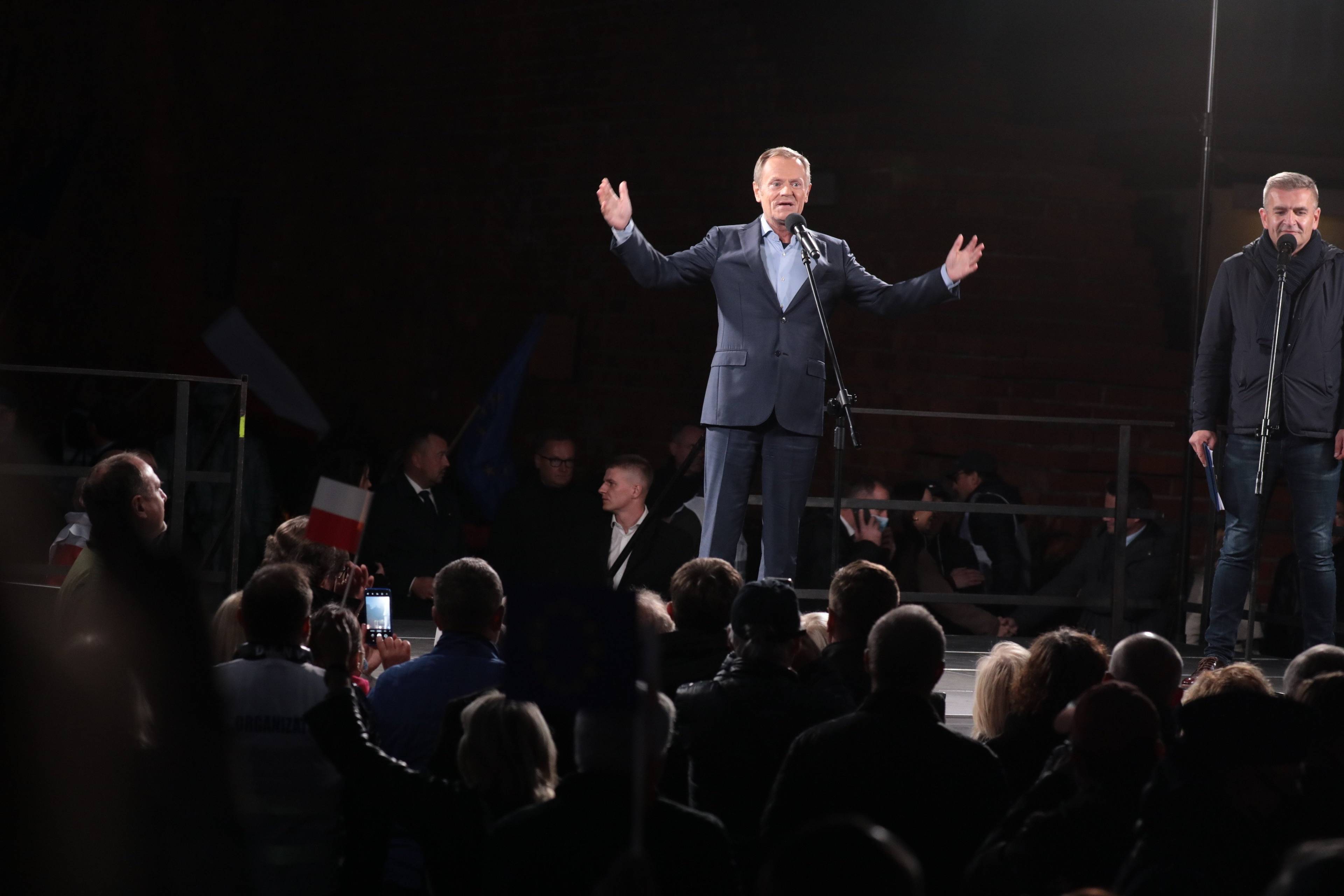 Mężczyzna w garniturze stoi na scenie z rozłożymi w bok rękami, przed nim w półmroku stoi tłum ludzi