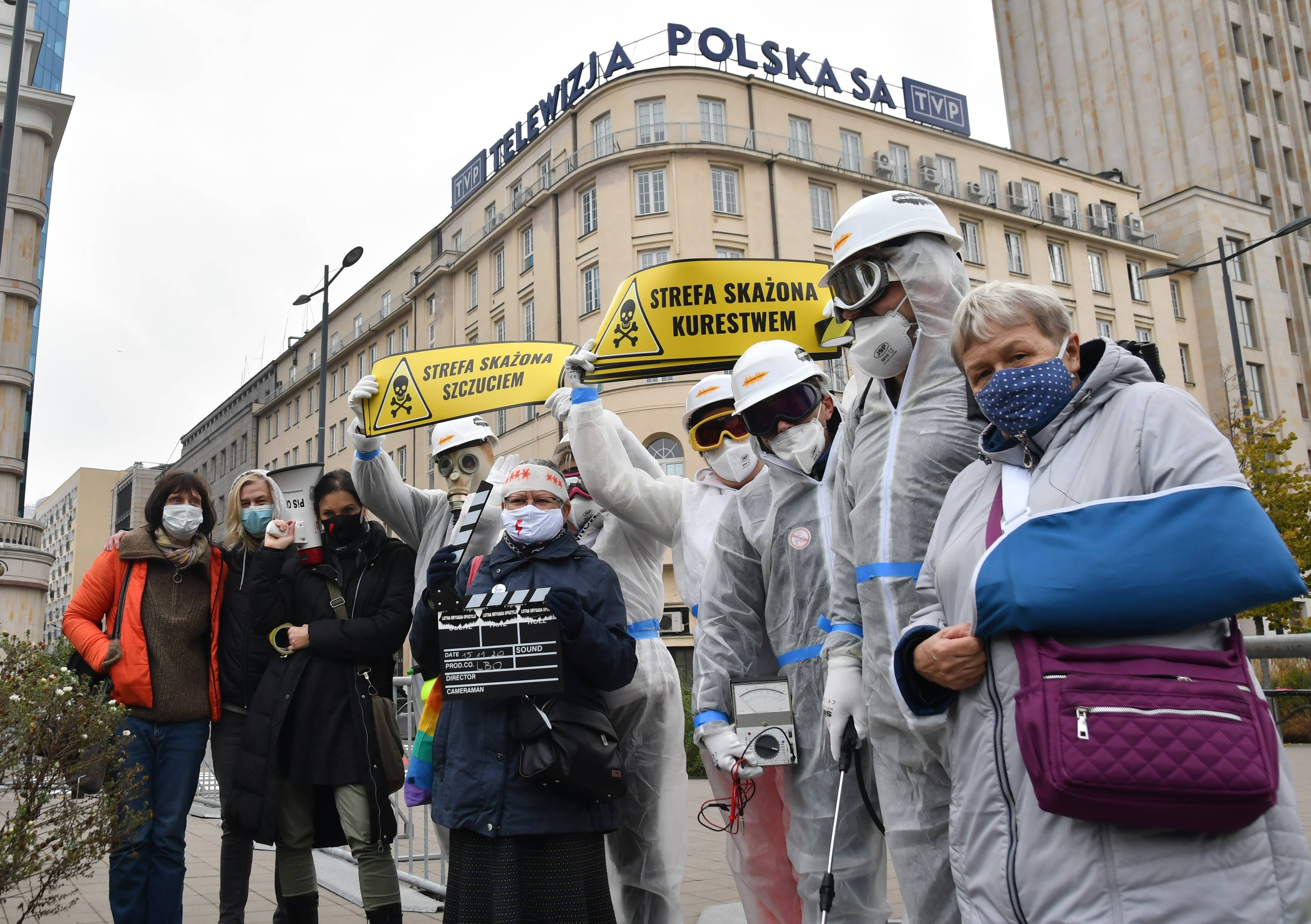 Lotna Brygada Opozycji pod TVP.Aktywiści w maskach przeciwgazowych i w kombinezonach stoją pod budynkiem TVP, za nimi żółte tabliczki ostrzegawcze