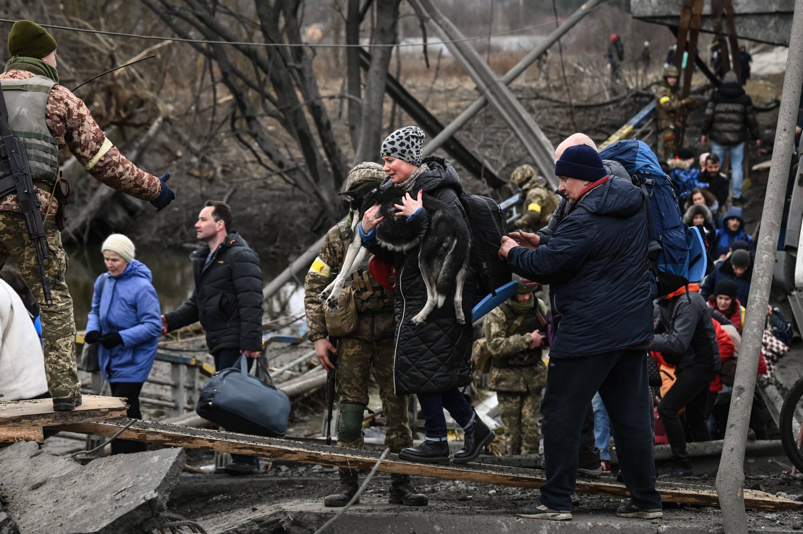 A woman carries a dog  while people cross a destroyed bridge as they  evacuate the city of Irpin, northwest of Kyiv, during heavy shelling and bombing on March 5, 2022, 10 days after Russia launched a military in vasion on Ukraine. (Photo by Aris Messinis / AFP)
