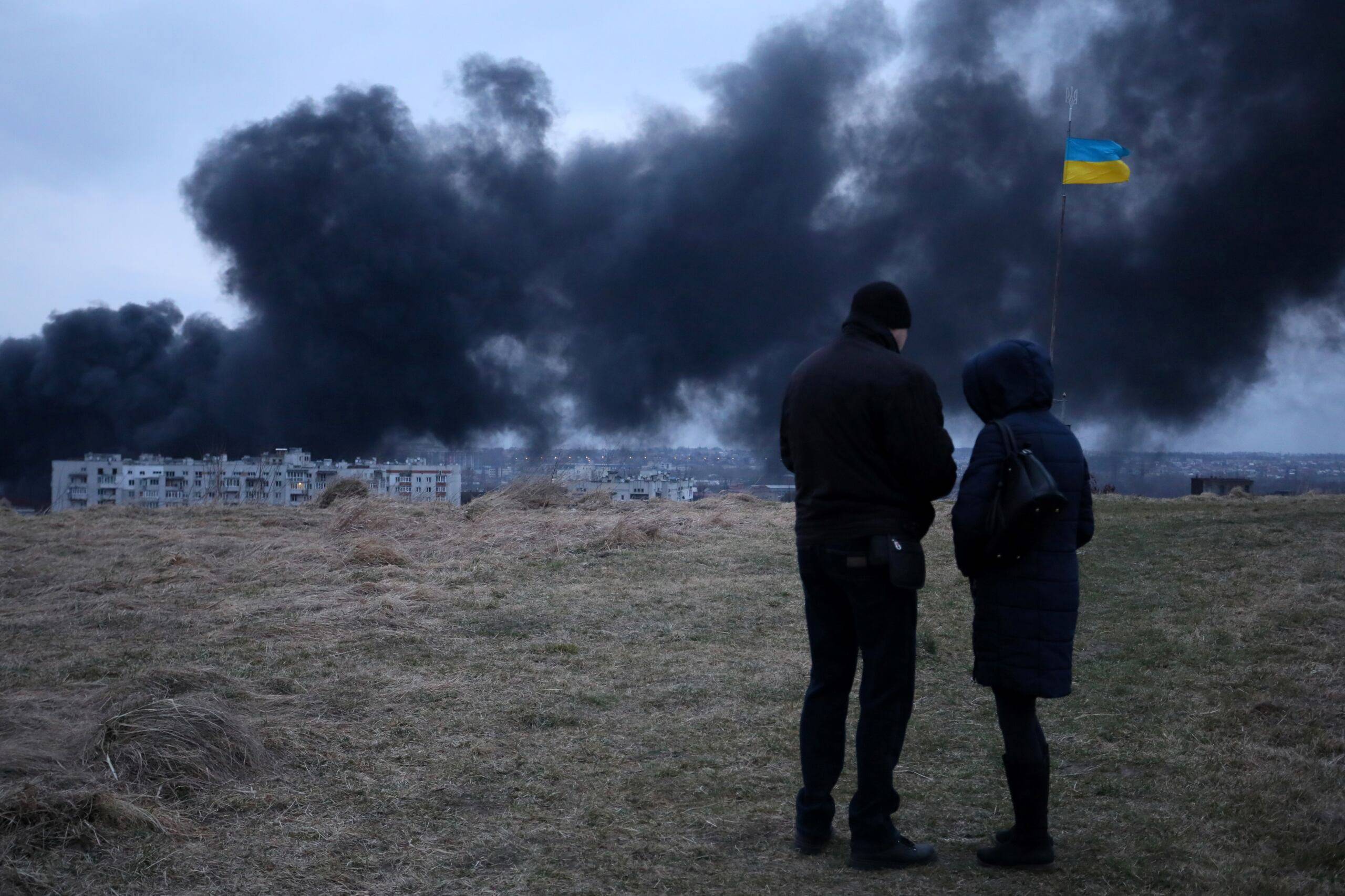 Ukraina, Lwów, 26.03.2022. Dym z płonących magazynów paliwa. Na pierwszym planie dwie postaci i flaga Ukrainy