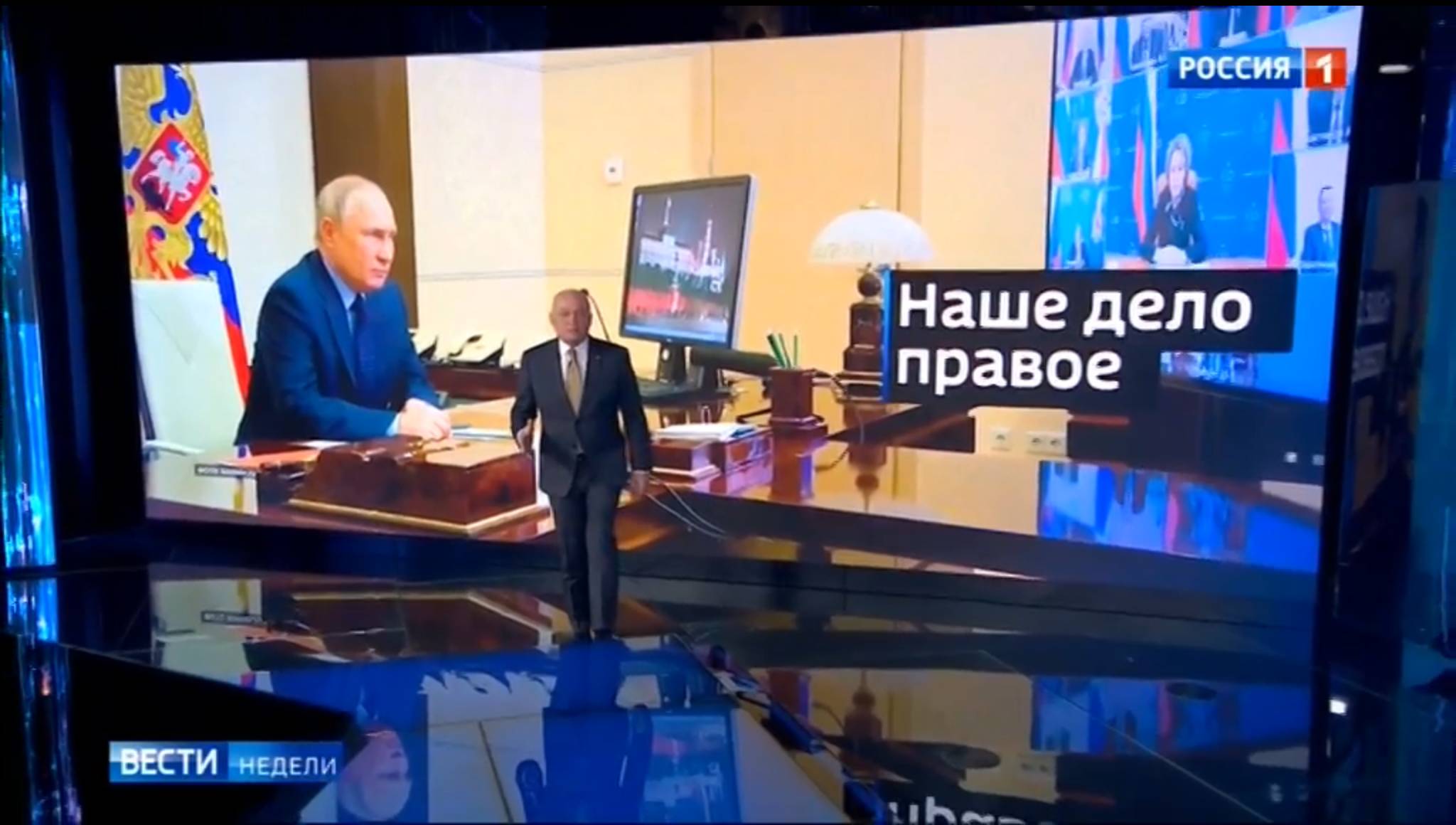 Studio telewizyjne. Prowadzący na pierwszym planie, w tle - zdjęcie Putina prowadzącego telekonferencję