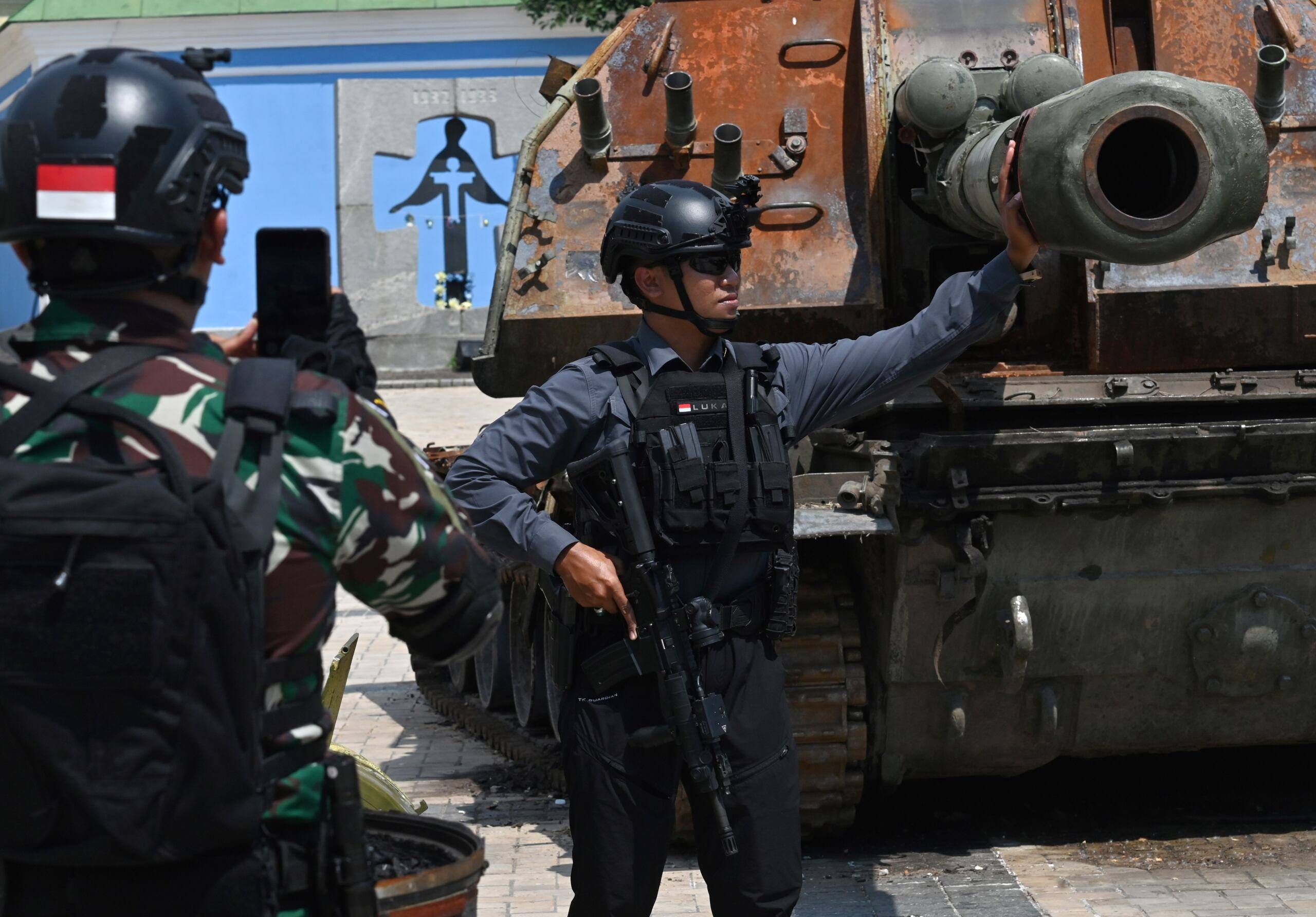 Dwa uzbrojeni mężczyźni z czerwobo białym - indonezyjskimi - naszywkami pozują przed wrakiem rosyjskiej armaty