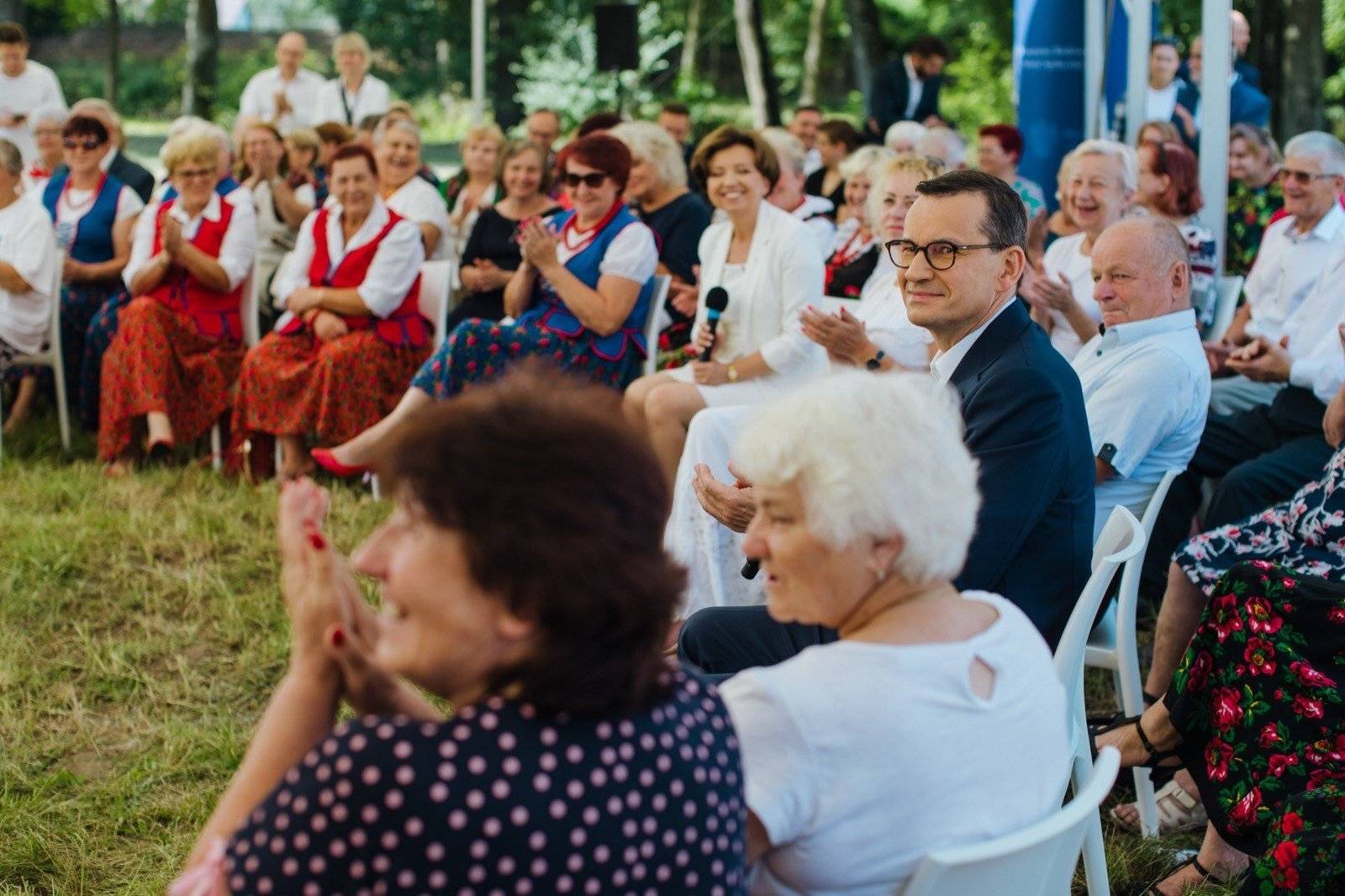 męzczyzna w okularach w średnim wieku (premier Morawiecki) w otoczeniu starszych kobiet siedzących w kręgu na trawie