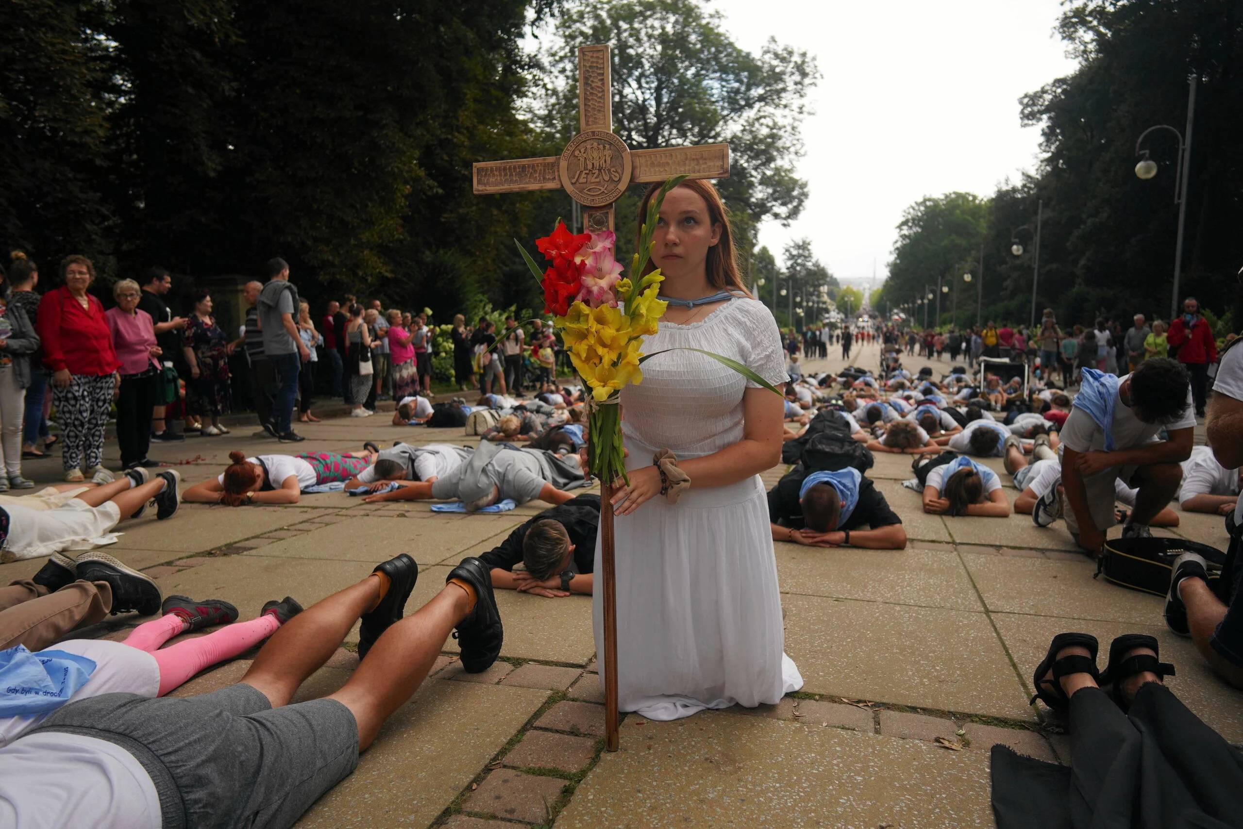 Kobieta w białej sukni klęczy trzymając wielki krzyż, wokół leżą na ziemi pielgrzymi