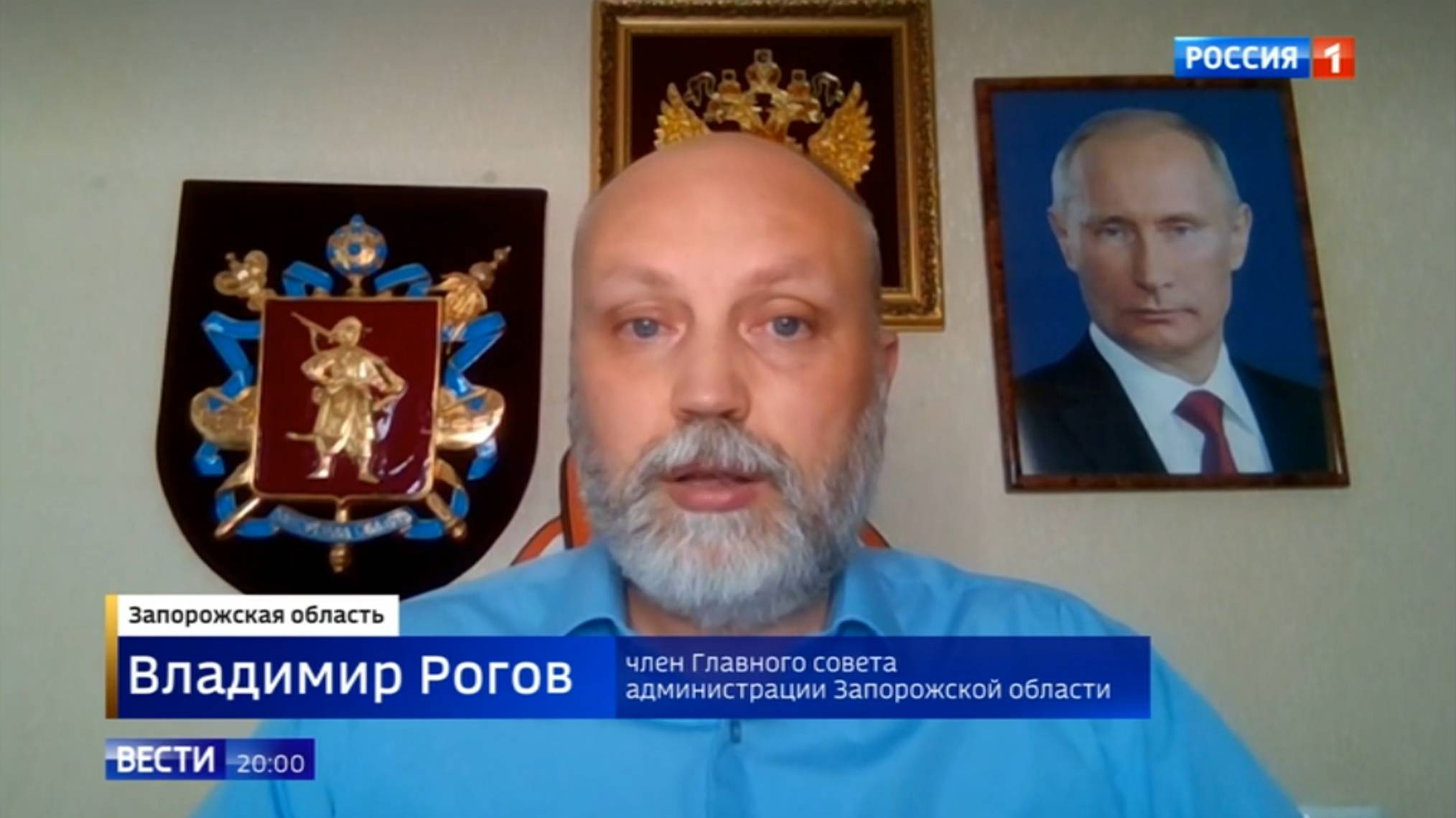 Mężczyzna w niebieskiej koszuli występuje na tle herbu Rosji i portretu Putina