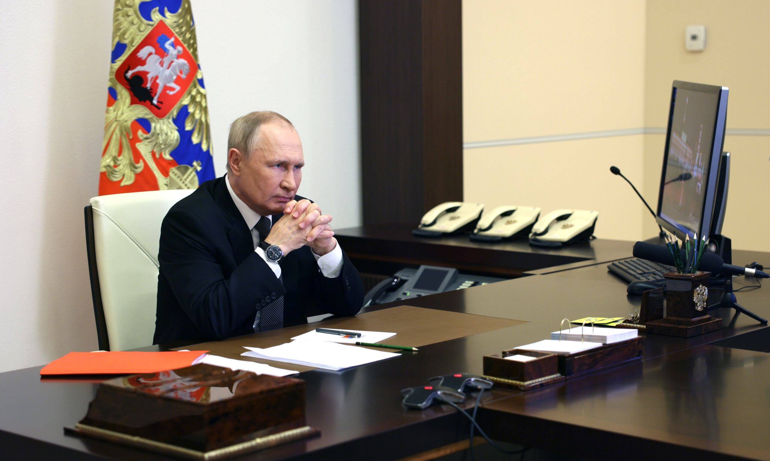 Władimir Putin siedzi za biurkiem, opiera o nie łokcie, dłonie ma splecione, wokół niego cztery aparaty telefoniczne