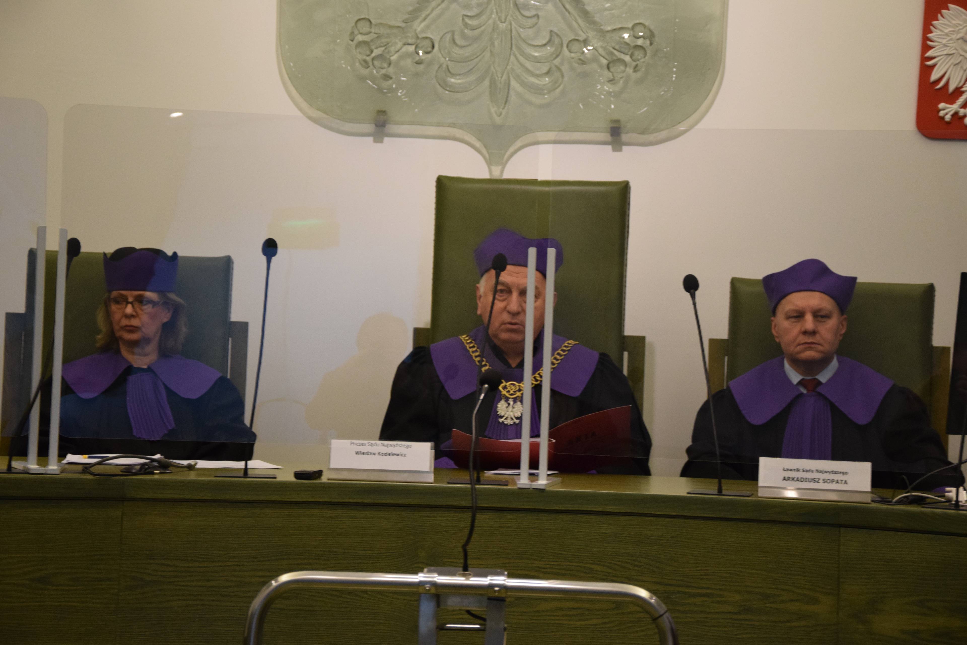 Trzej sędziowie w togach Sądu Najwyższego (fioletowe wyłogi i czapki) siedzą za stołem