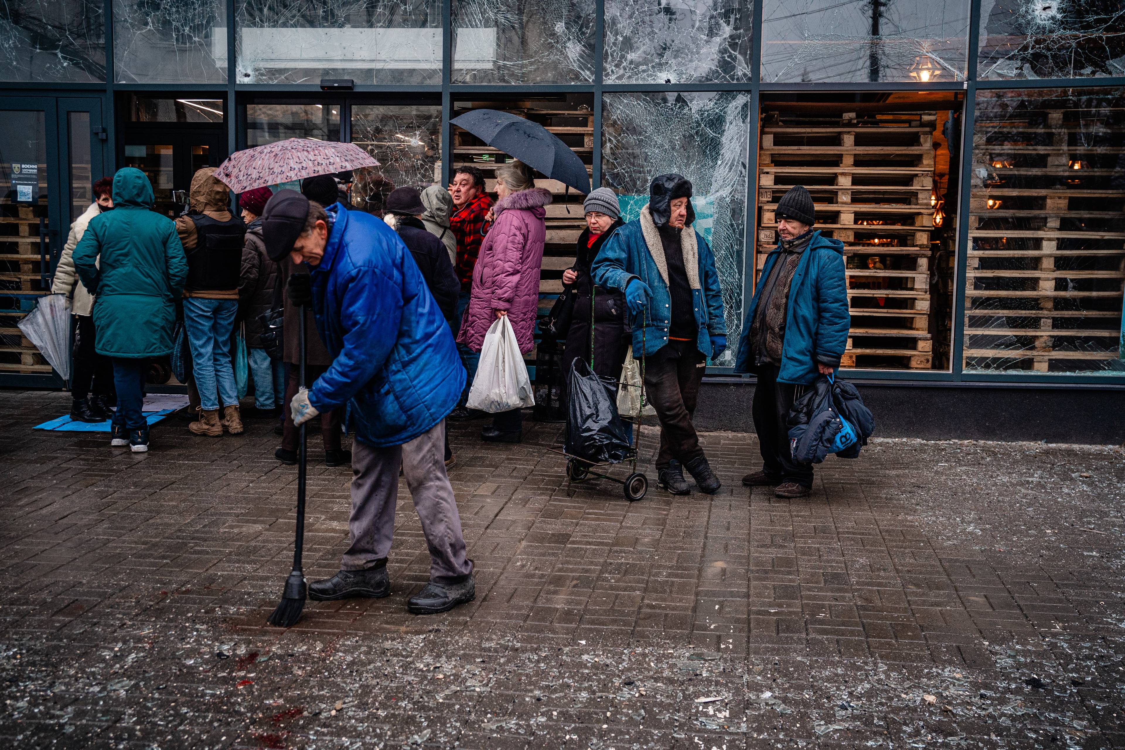 People stand in line outside a supermarket while a man cleans up after shelling on Ukrainian city of Kherson on December 25, 2022, amid the Russian invasion of Ukraine. - Ukrainian President Volodymyr Zelensky blasted Russian "terror" after shelling left 10 dead and 55 injured in Kherson city on December 24, 2022, urging his compatriots to persevere as they observed a Christmas Eve defined by war. (Photo by Dimitar DILKOFF / AFP)