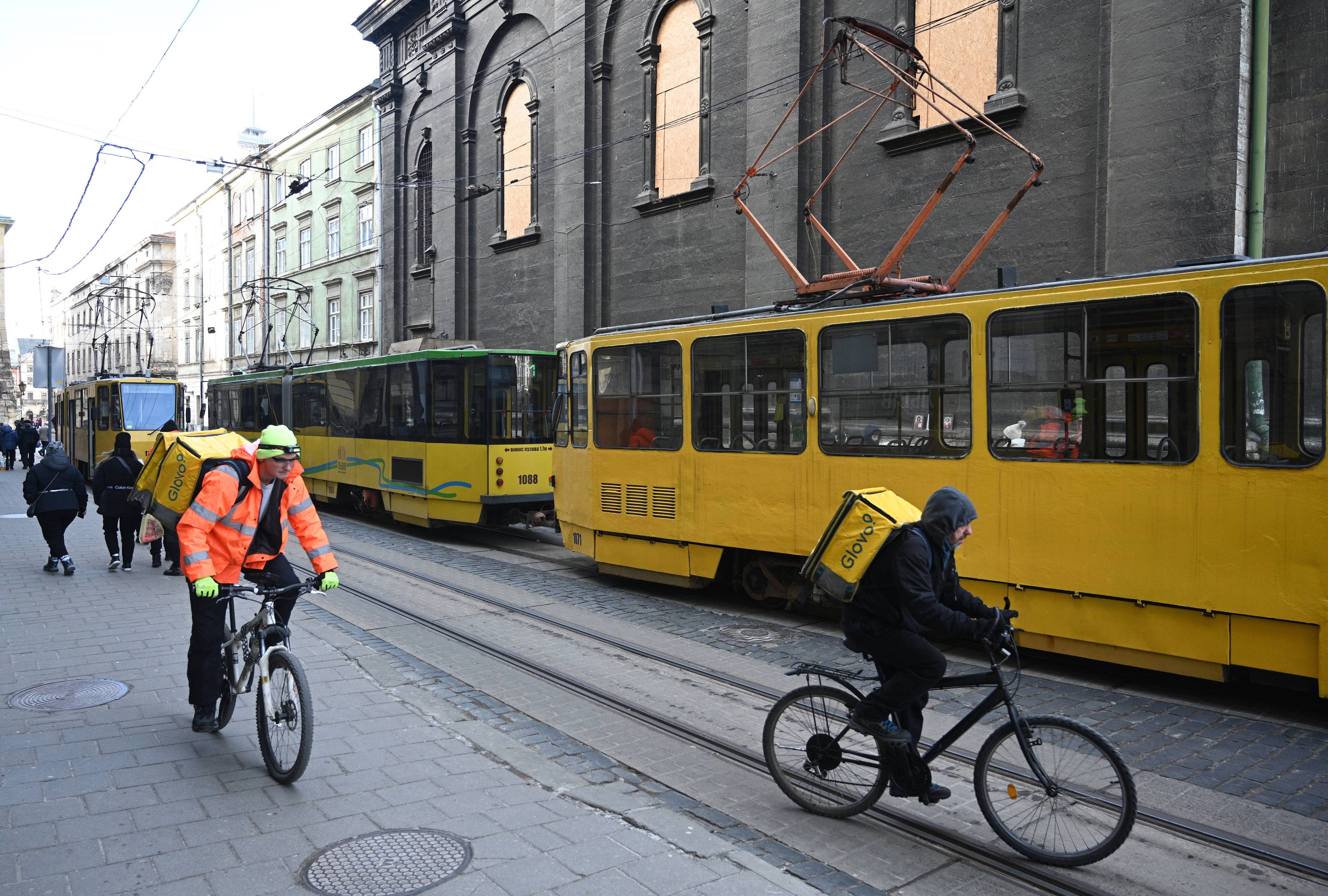 Na wąskiej staromiejskiej ulicy stoją puste żółte tramwaje. Ulicą jadą rowerzyści z ładunkami