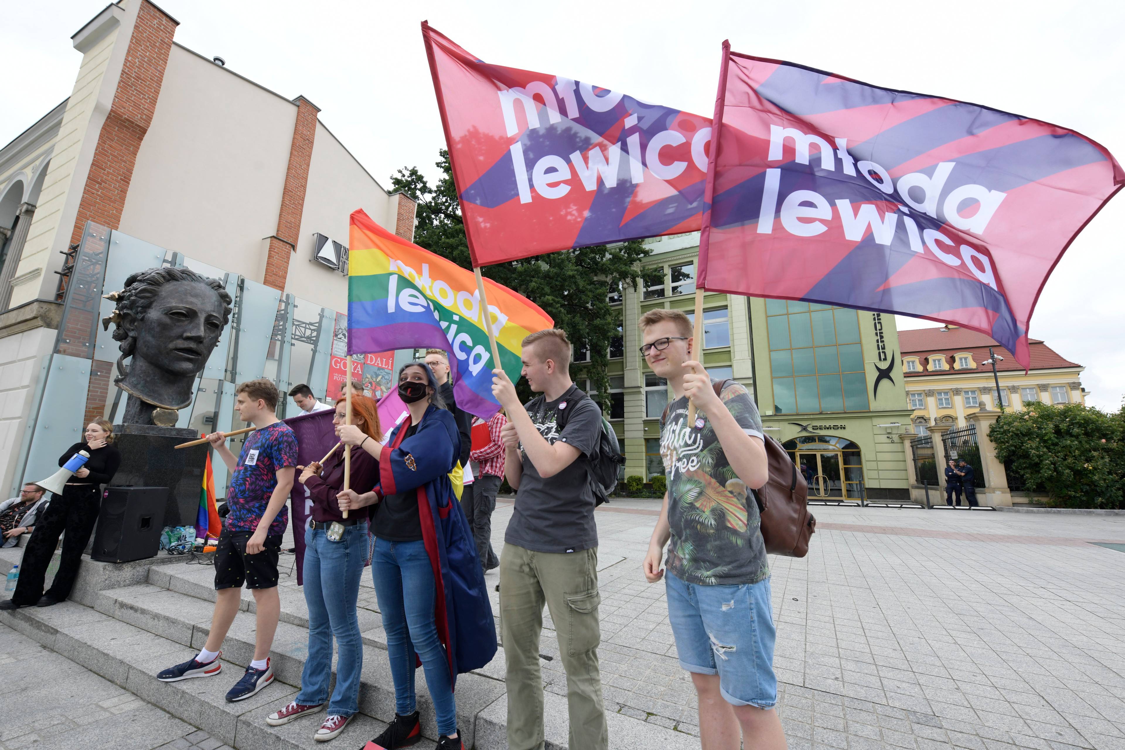 grupa młodzieży stoi na placu z powiewającymi flagami, na których jest napisa "Młoda lewica". Jena z flag jest w kolorach tęczy.