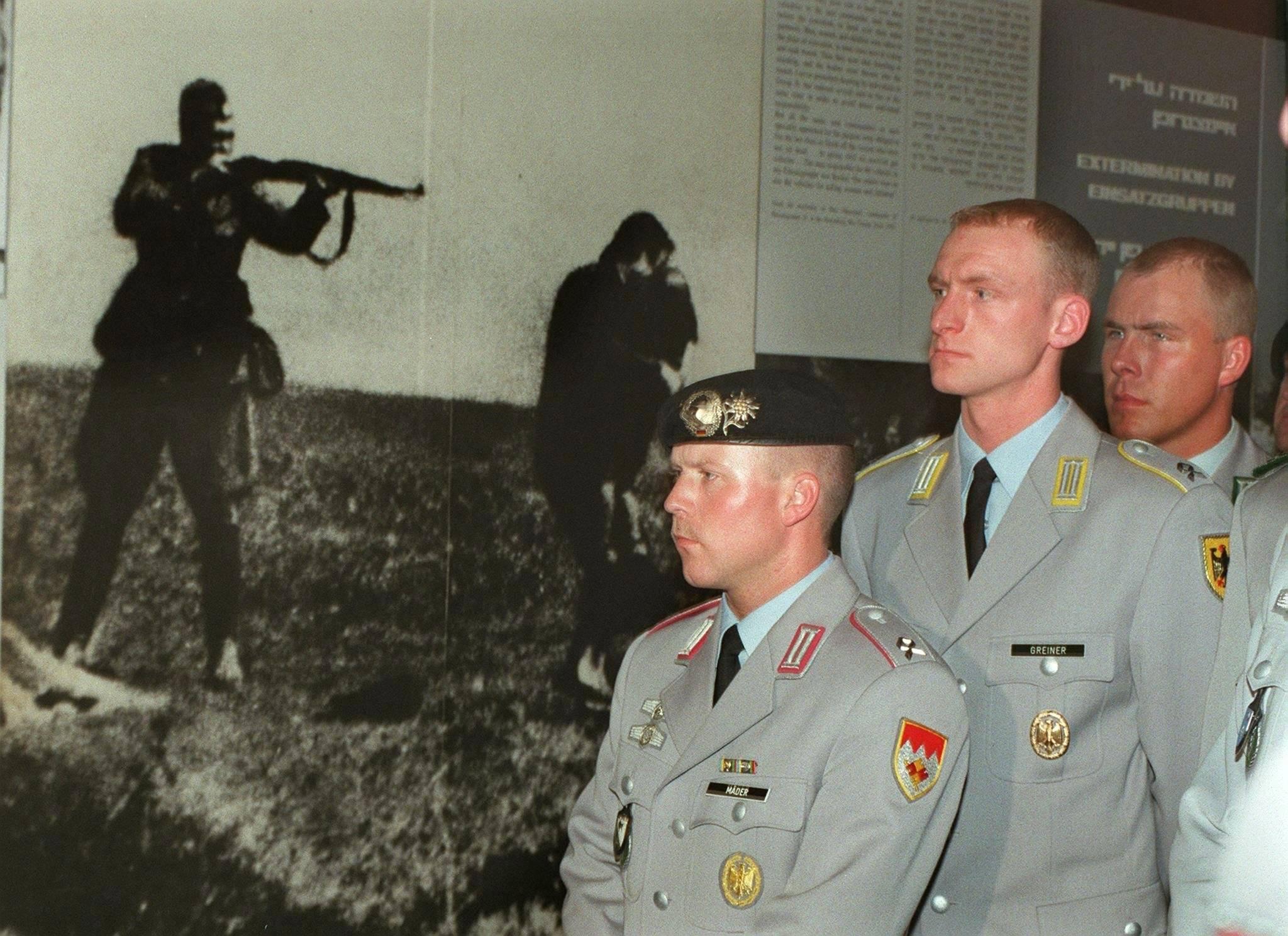 trzech oficerów Bundeswehry stoi na tle zdjecia przedstawiającego żołnoerza Wehrmachtu, strzelającego do kobiety z dzieckiem