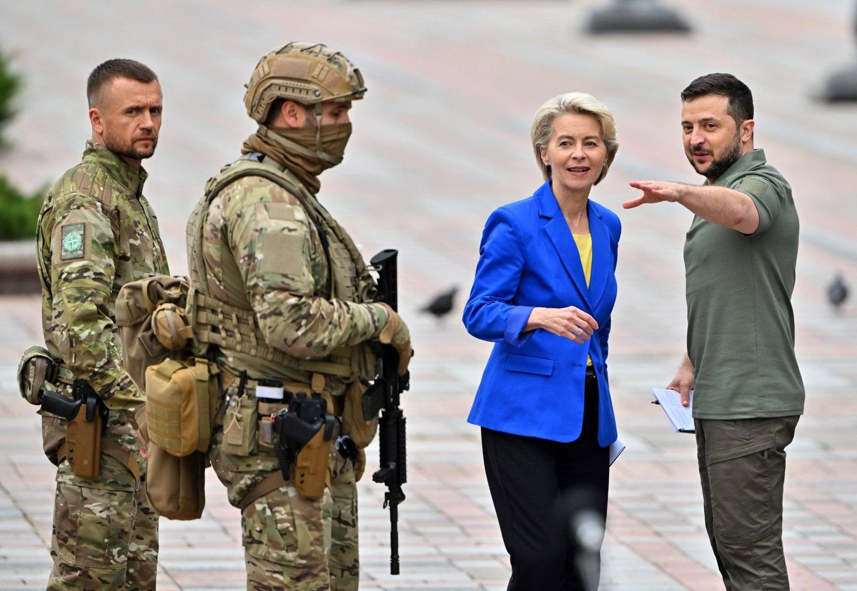 Dwaj uzbrojeni żołnierze obok przewodniczącej KE Ursuli von der Leyeen w niebieksikm żakieci i prezydenta Zełenskiego w wojskowym ubraniu