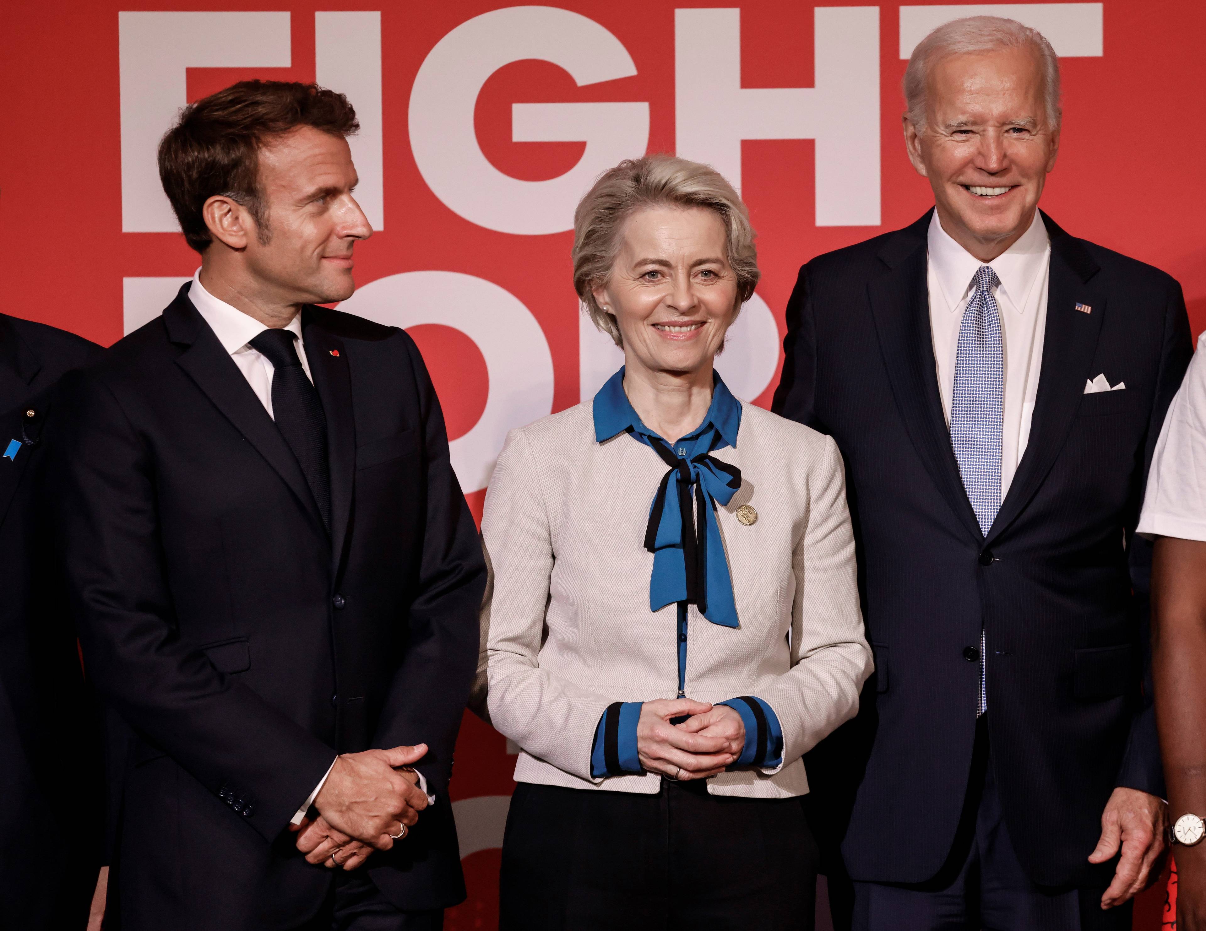 Obok siebie na czerwonym tle stoi troje przywódców, uśmiechają się: od lewej prezydent Francji Emmanuel Macron, szefowa Komisji Europejskiej Ursula von der Leyen i prezydent Stanów Zjednoczonych Joe Biden