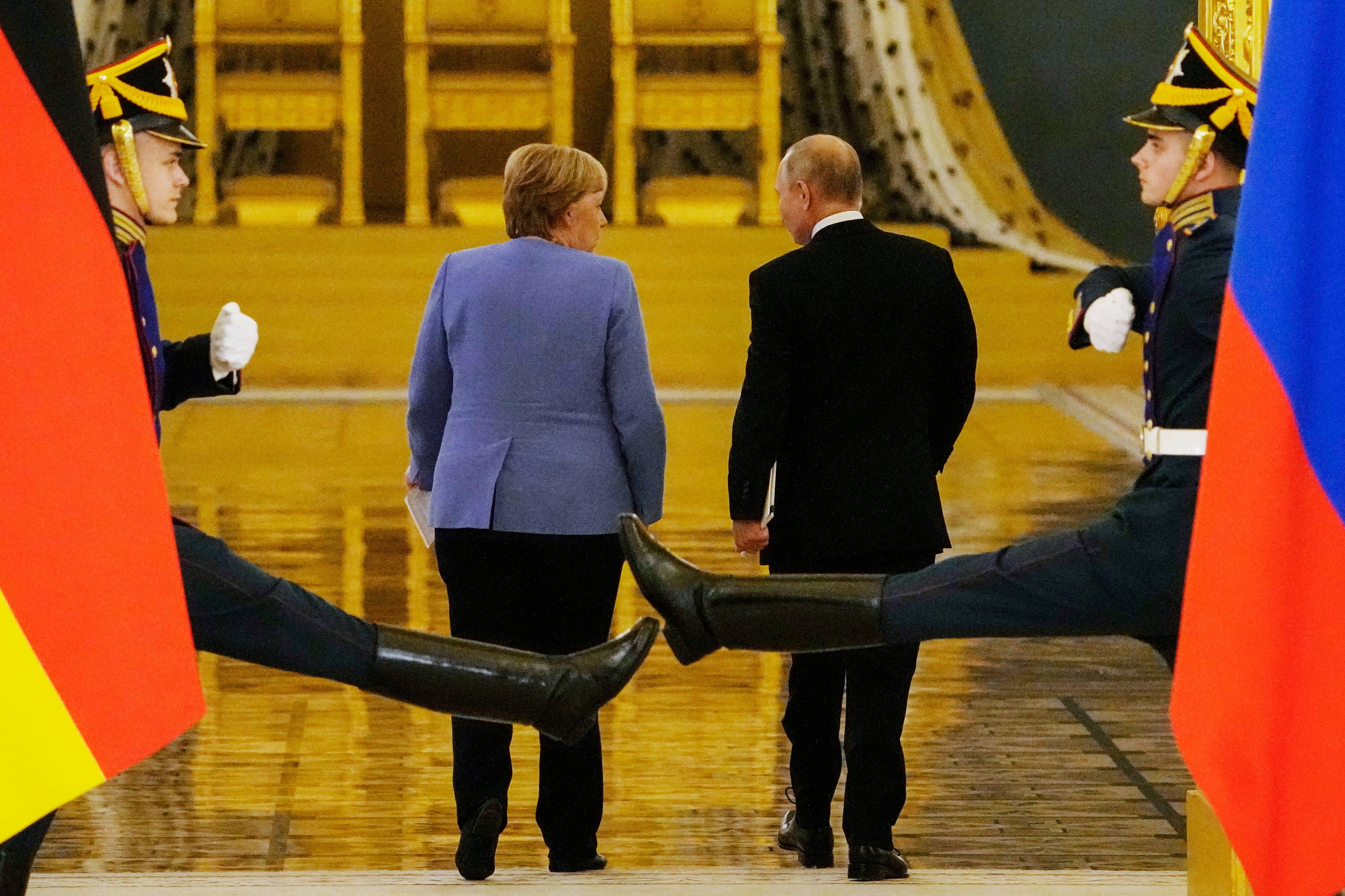 Putin i Merkel sfotografowani od tyłu idą przez wielką salę na Kremlu. Na pierwszym planie - nogi paradnej rosyjskiej gwardii w wykopie. Ich nogi wyprostowane są na wysokości pośladków dwojga dostojników